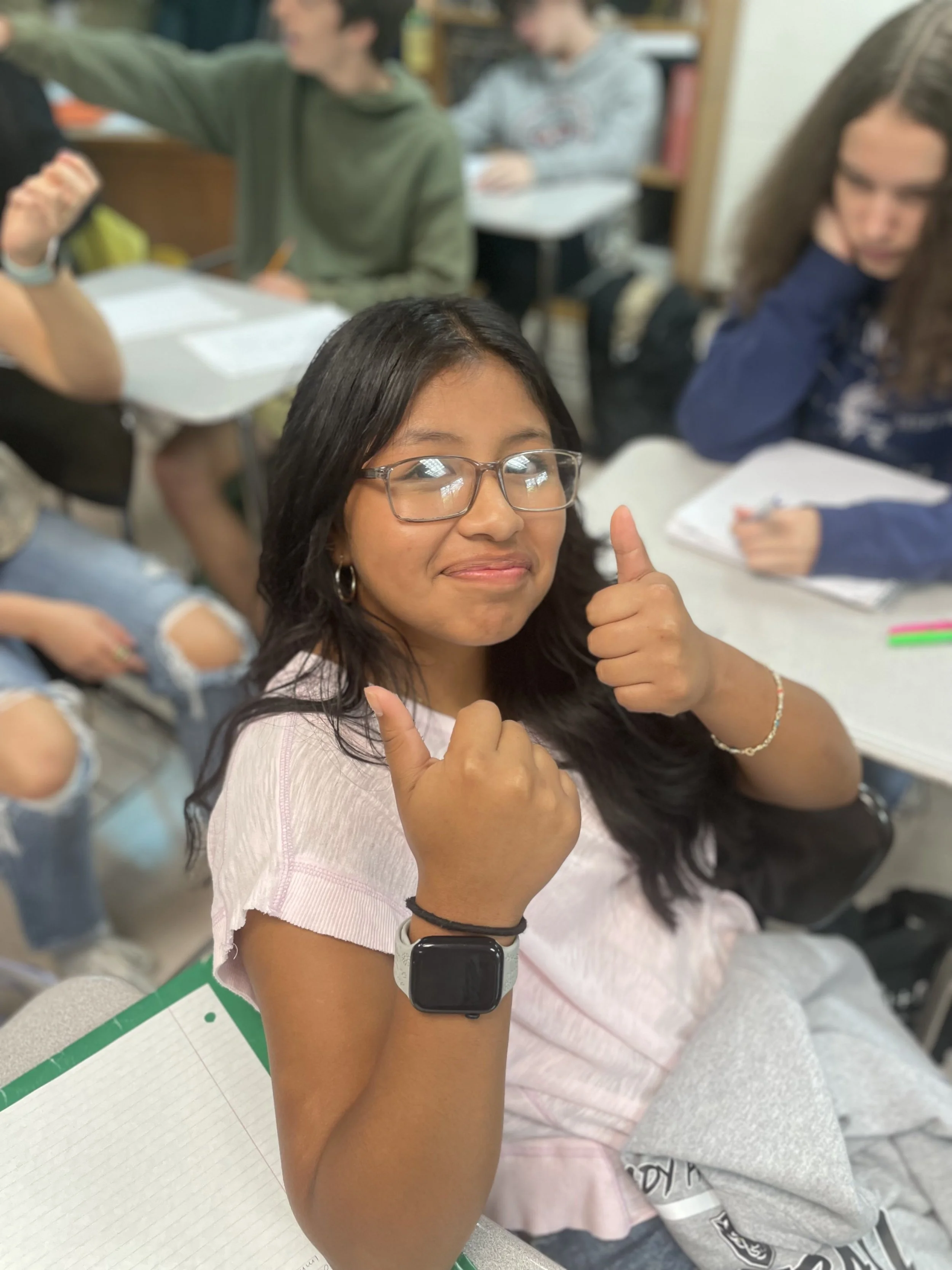 A girl wearing glasses showing a thumbs-up gesture on her right hand, sitting in a classroom with other students in the background.