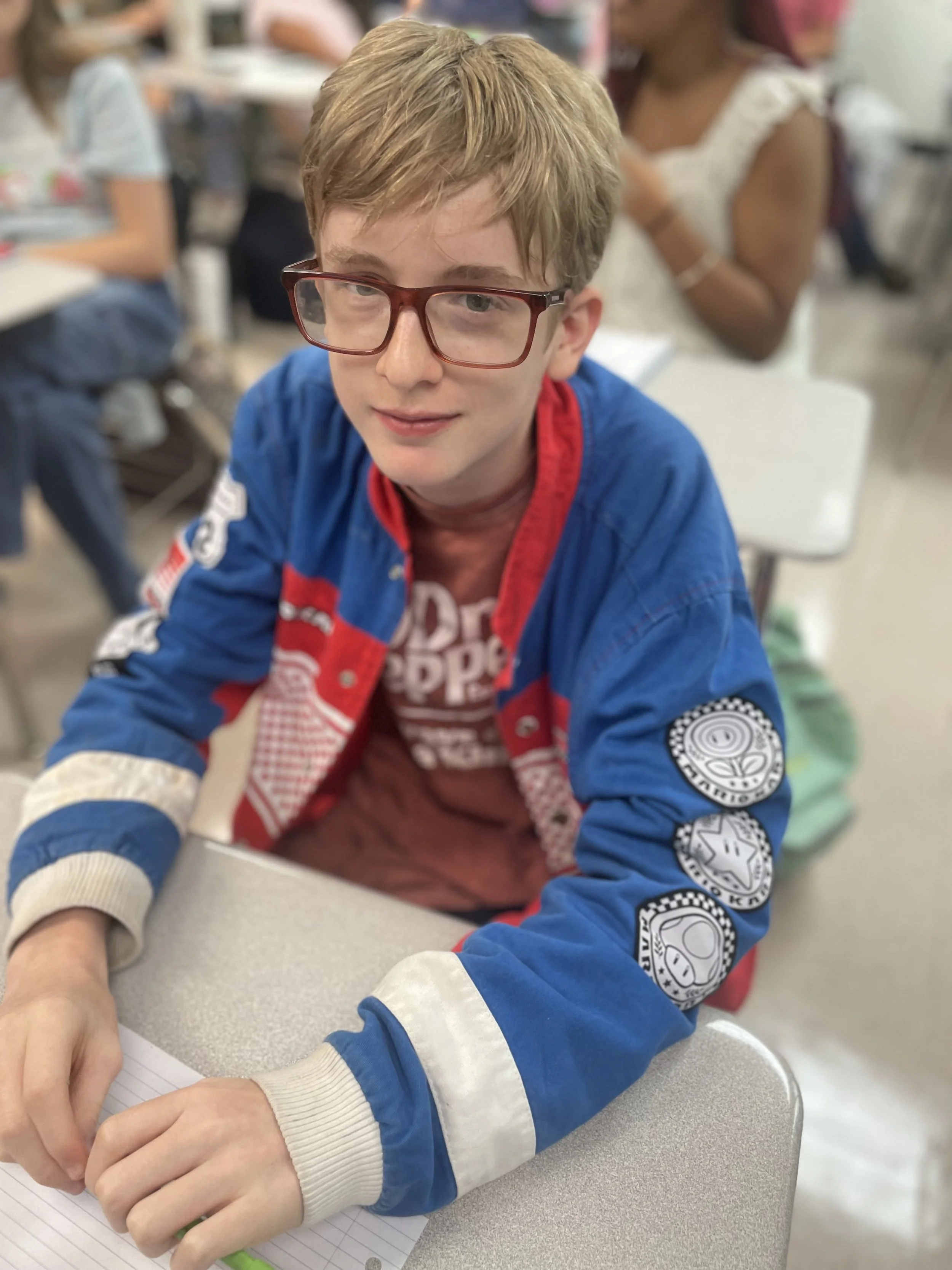 A young boy with red glasses and light brown hair wearing a blue and red jacket sits at a desk in a classroom, with other students in the background.