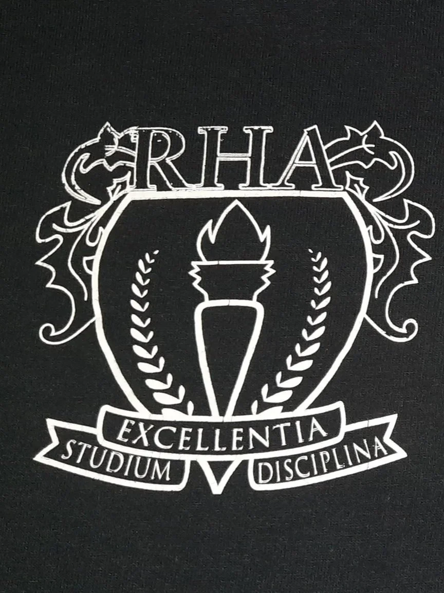 A black shirt with a white emblem featuring a torch, laurel branches, and banners displaying the words 'ARHAE' at the top, 'EXCELLENTIA' in the middle, and 'STUDIUM DISCIPLINA' at the bottom.