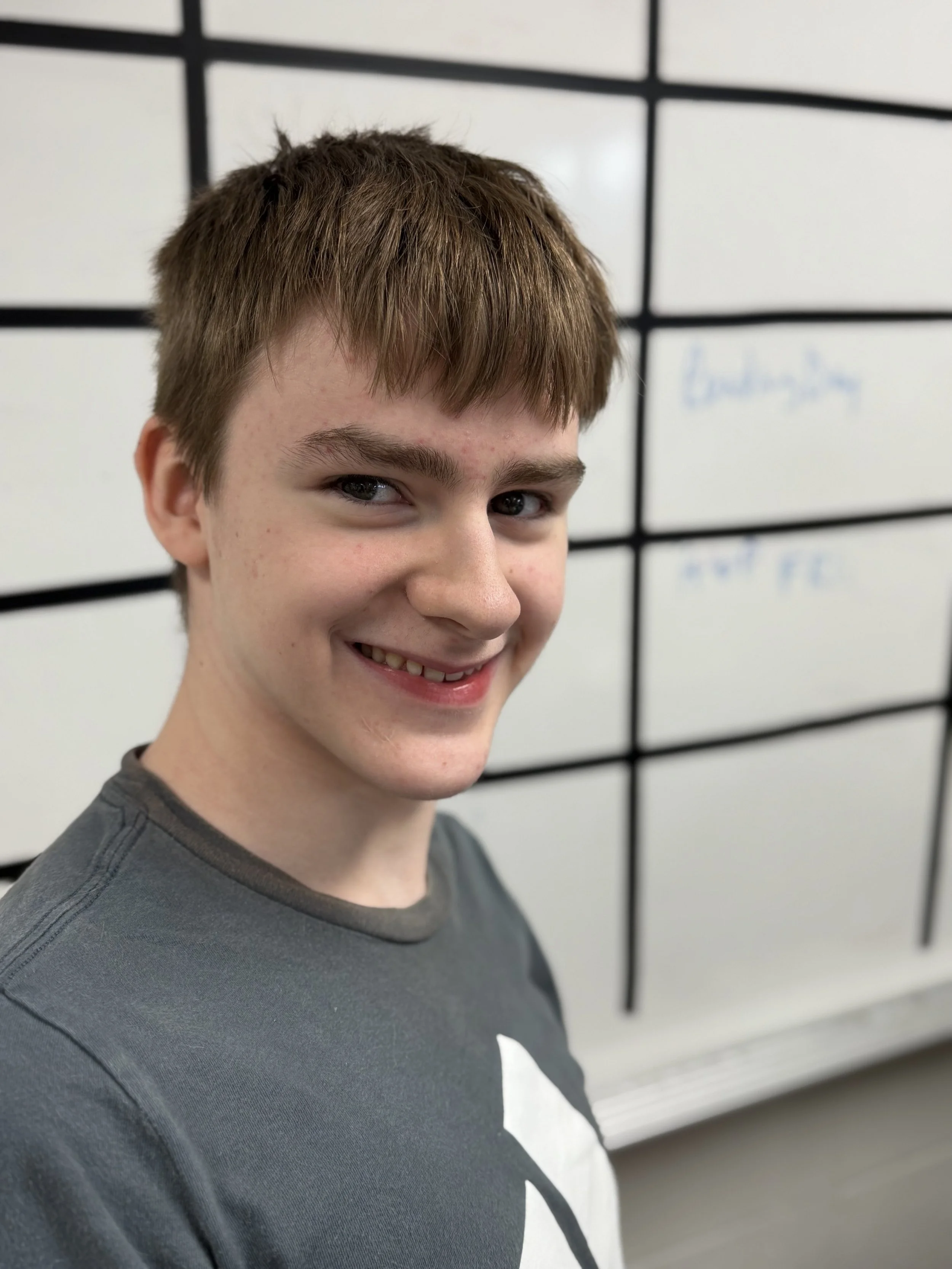 A smiling young boy with short brown hair and light skin standing in front of a whiteboard.