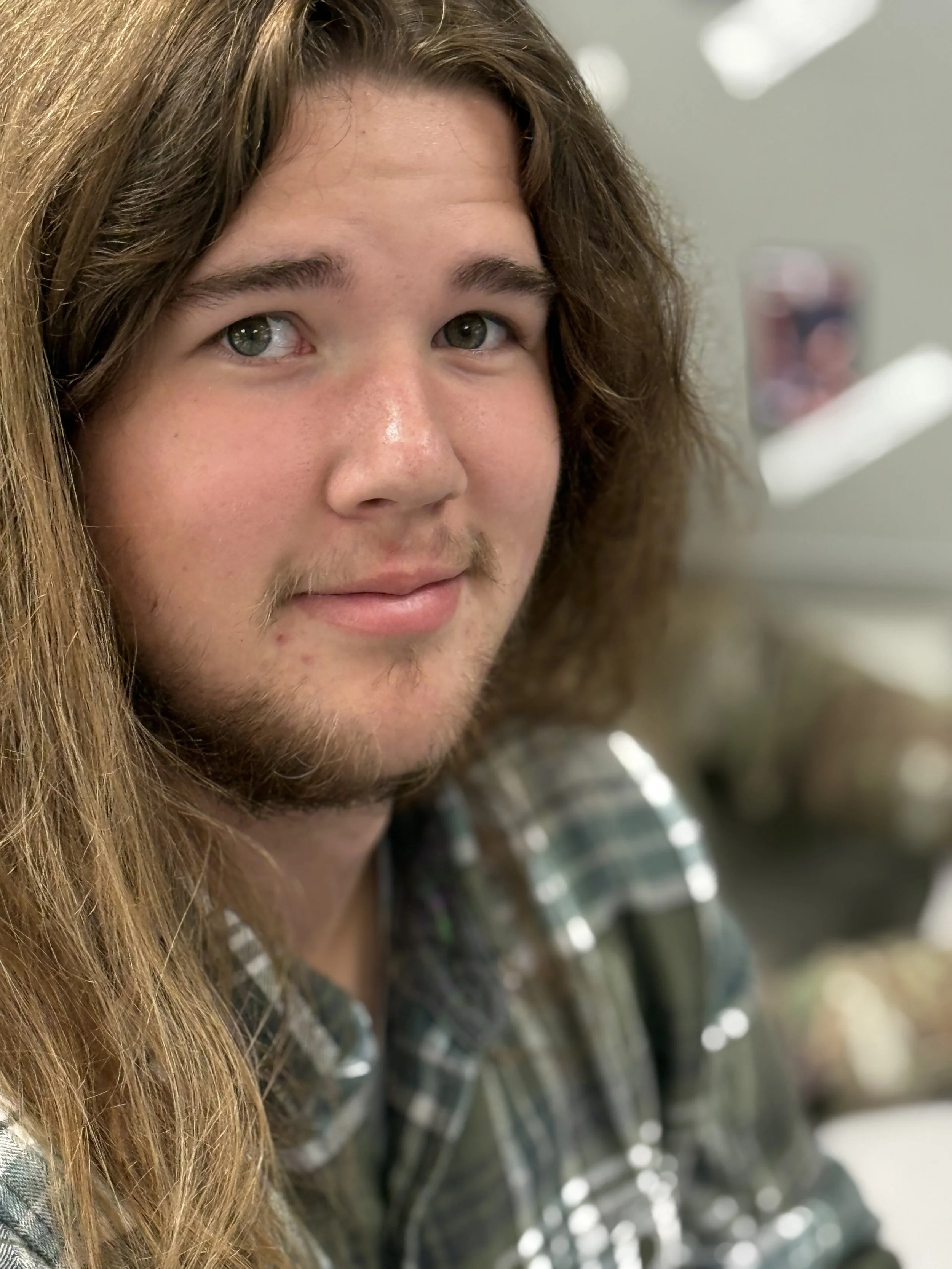 A young man with long hair and a slight beard wearing a plaid shirt, looking slightly to the side with a subtle smile.