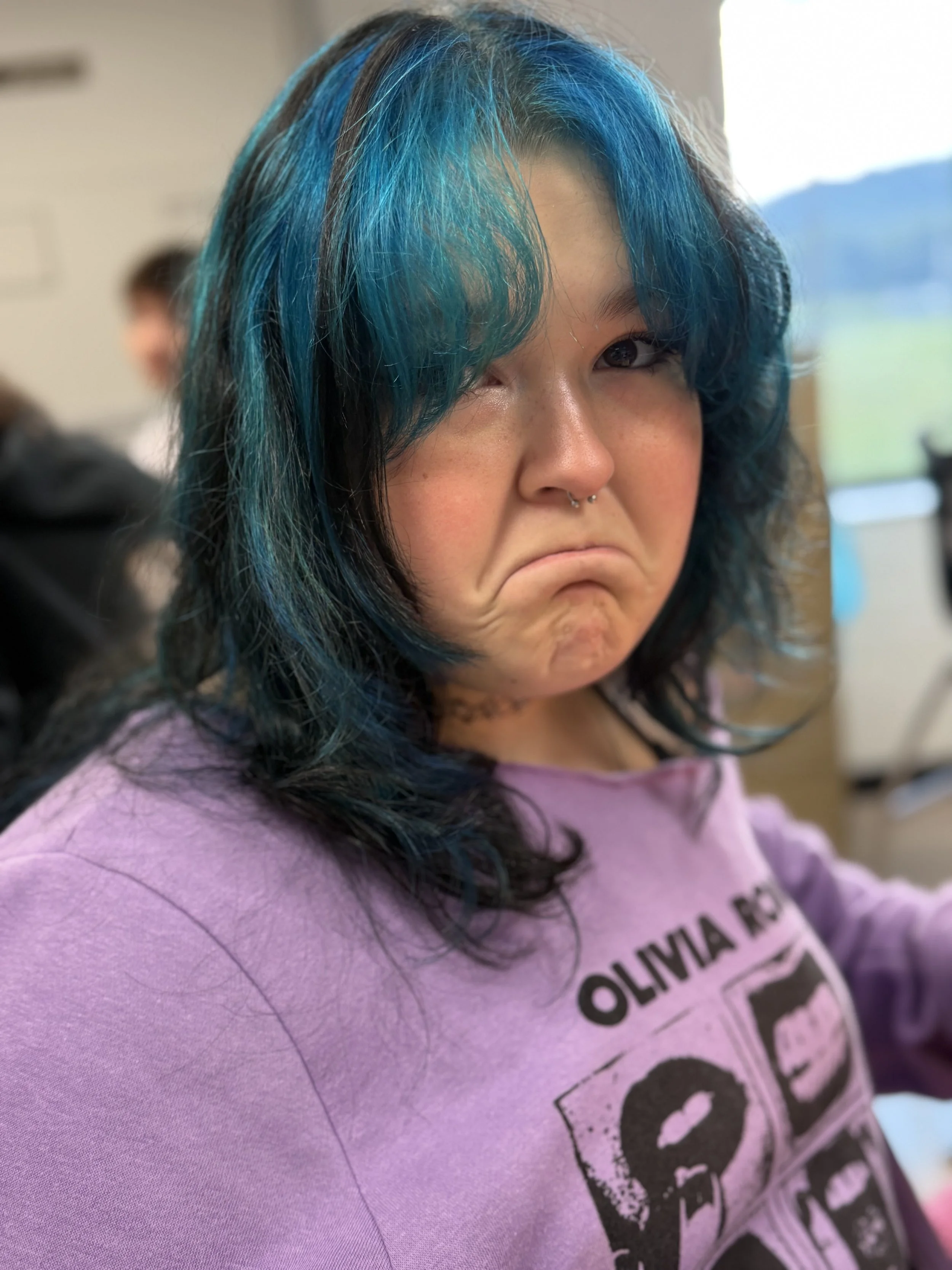 A young woman with black hair dyed blue at the tips, wearing a purple sweatshirt with a graphic design and the words 'Olivia' on it. She is making a sad or disappointed facial expression.