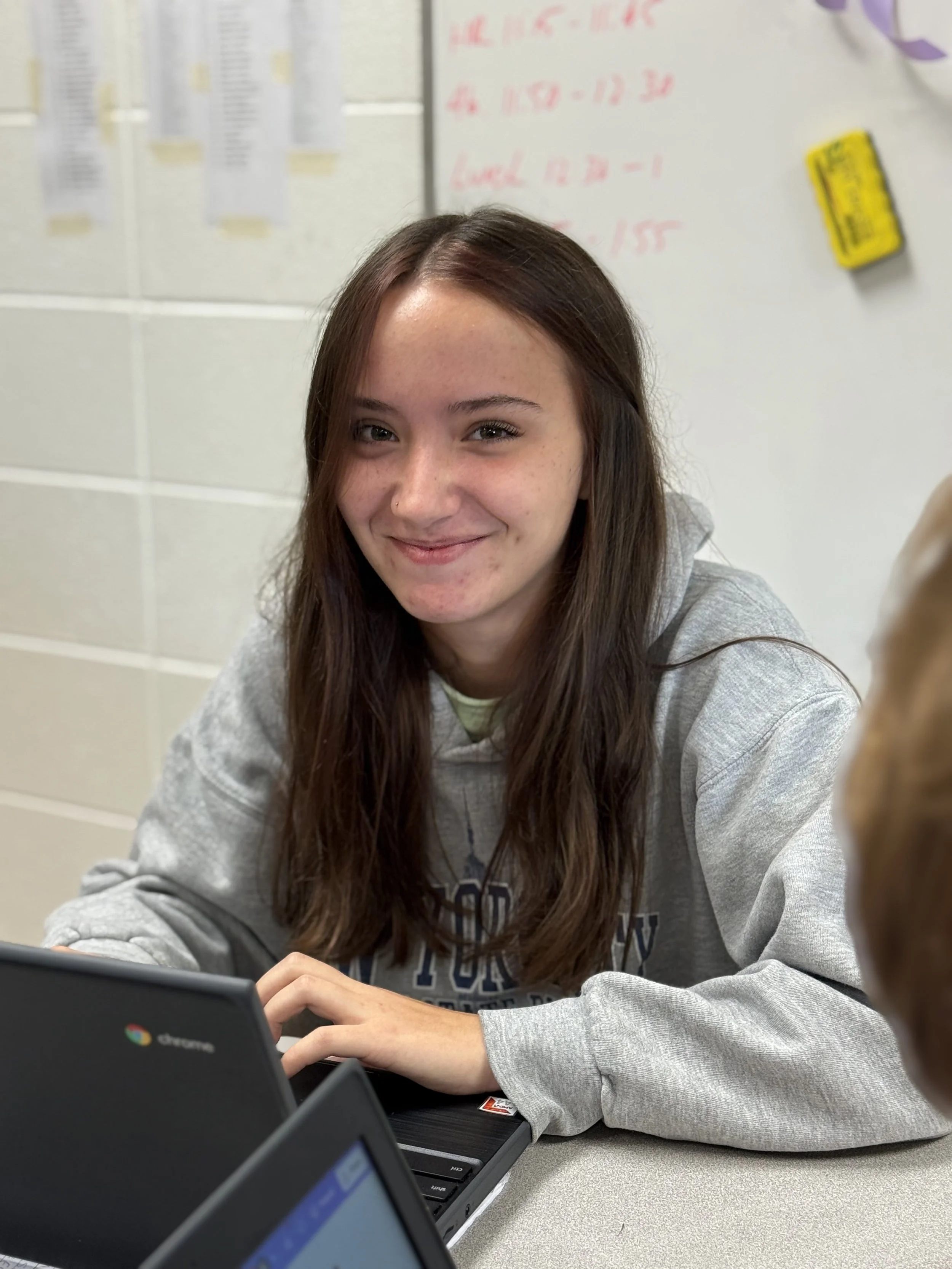A girl with long brown hair smiling while sitting at a table with laptops in a classroom setting.