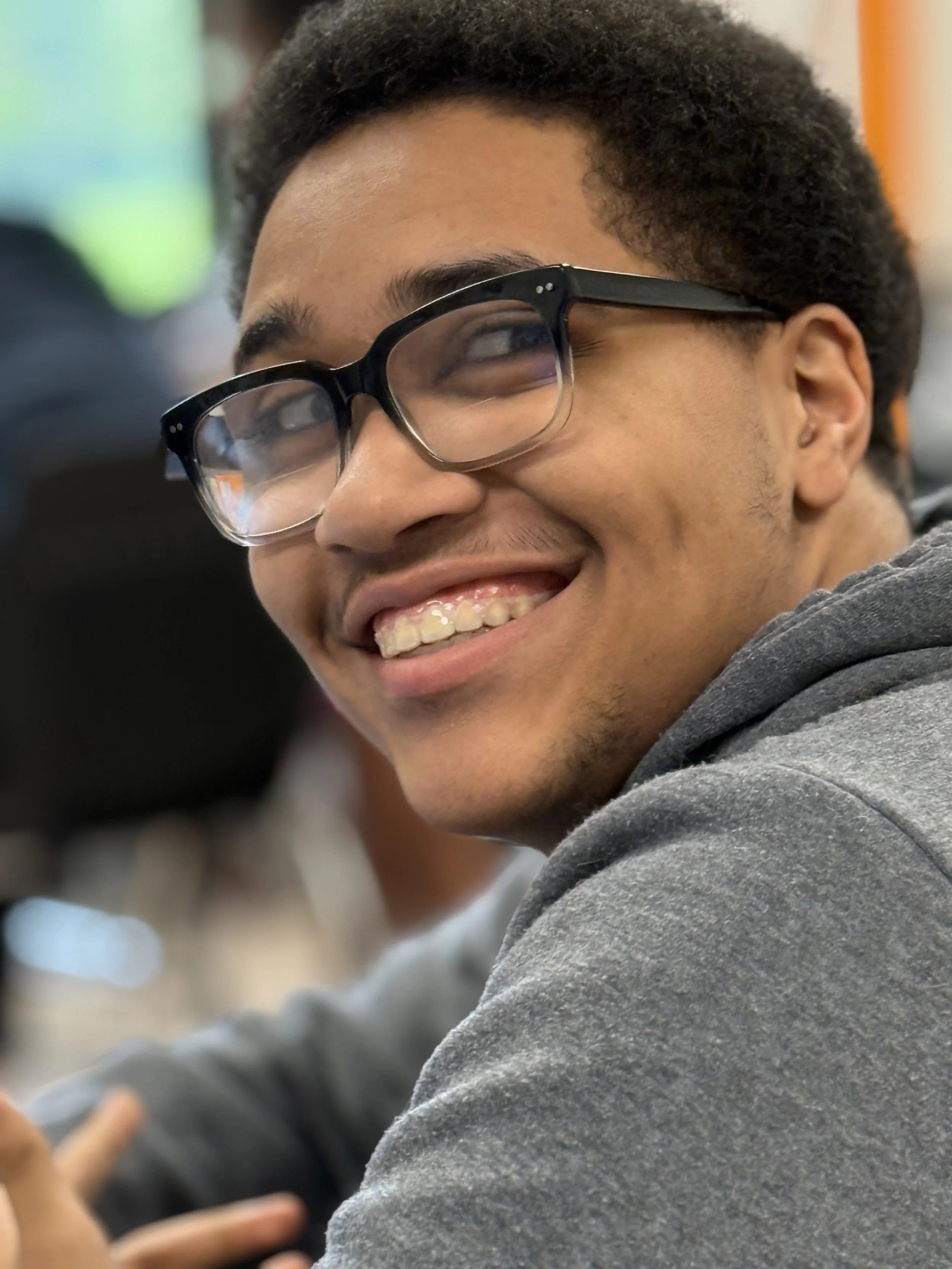 A young man with curly dark hair, wearing black glasses and a gray hoodie, smiling and looking over his shoulder in a classroom setting.