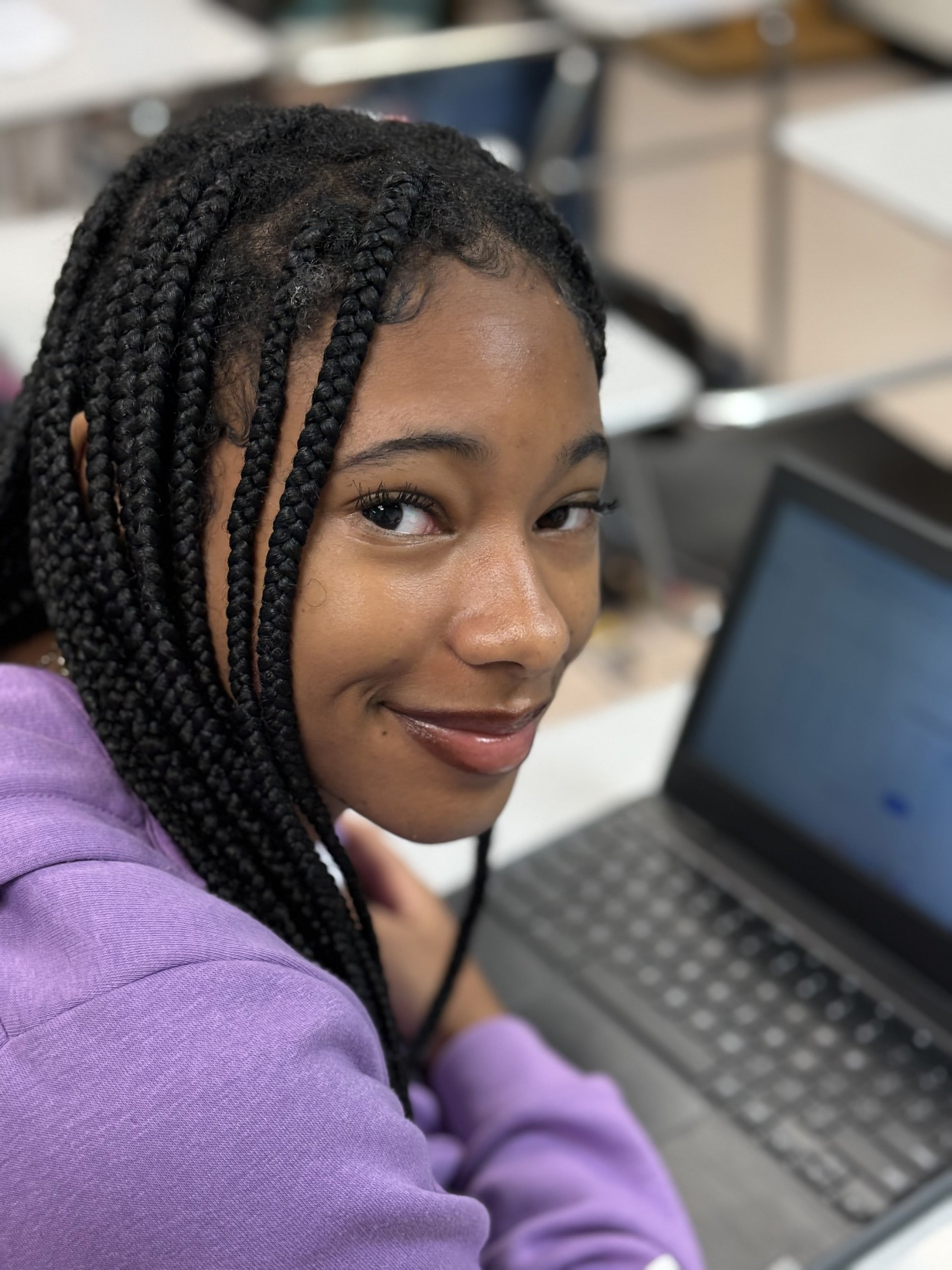 A young woman with braided hair wearing a purple sweatshirt sitting at a desk with a laptop.