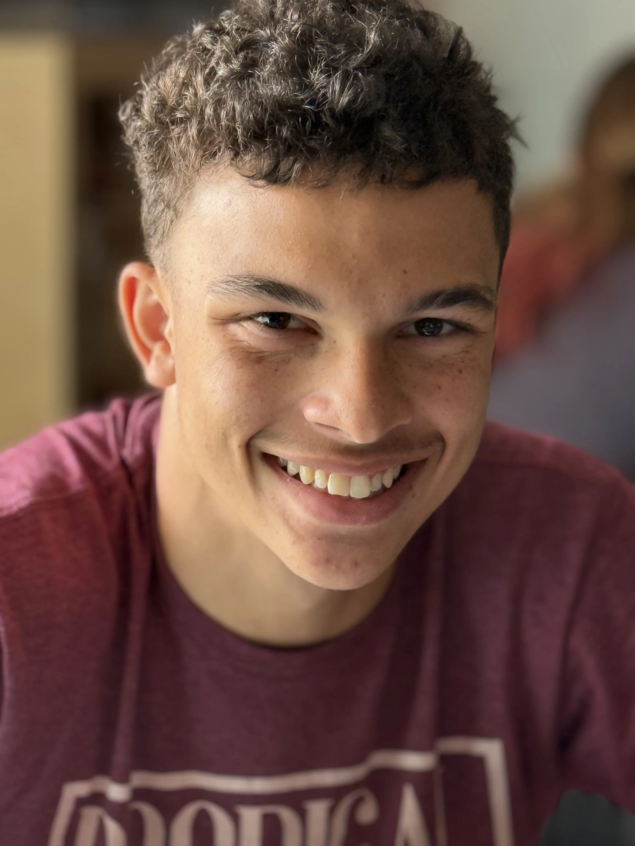 A smiling teenage boy with short, curly brown hair wearing a maroon t-shirt.