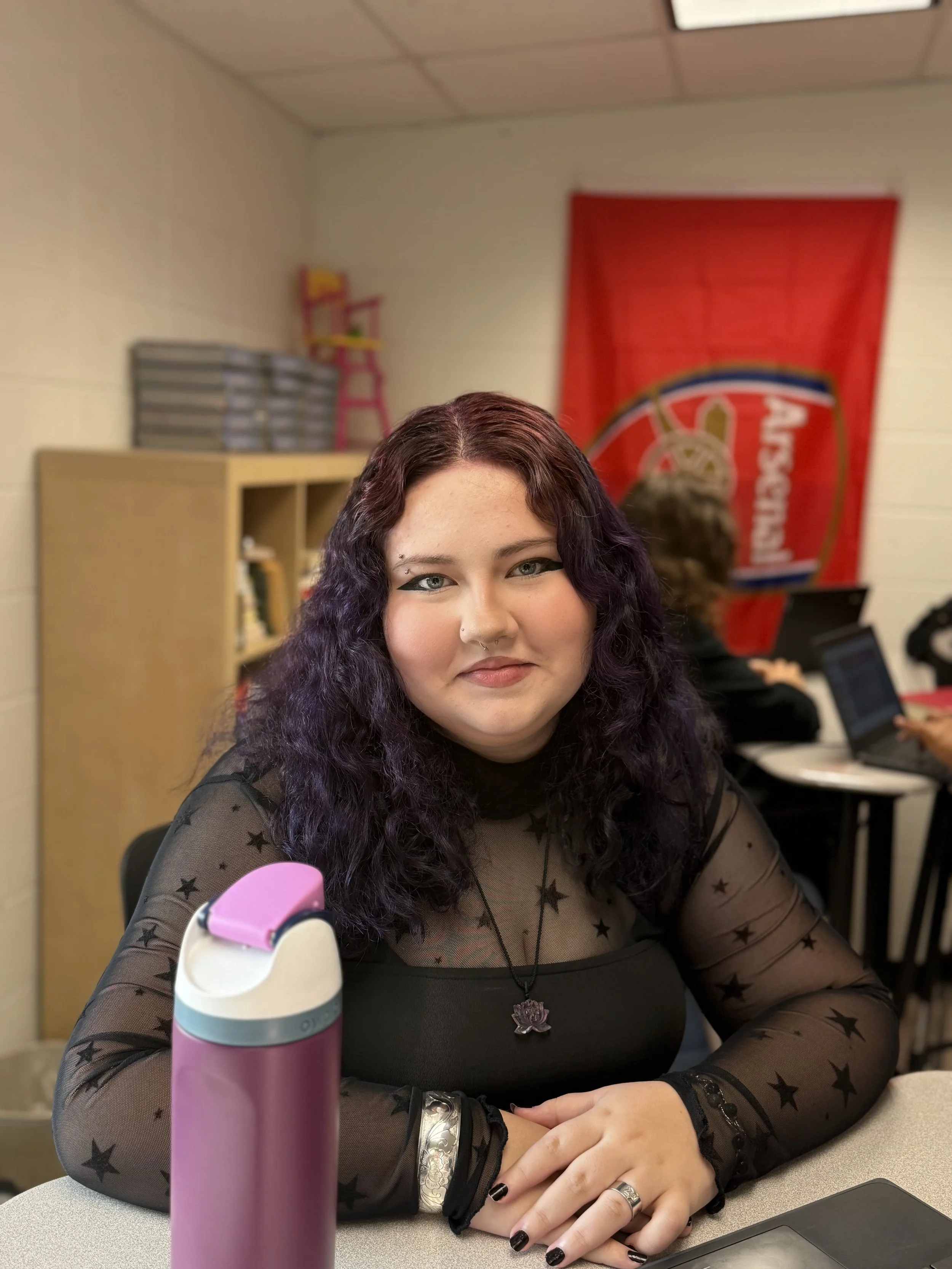 A young woman with dark purple curly hair sitting at a desk with a pink and white water bottle in the foreground, in a classroom or office setting, with a woman working on a laptop in the background and a red banner on the wall.