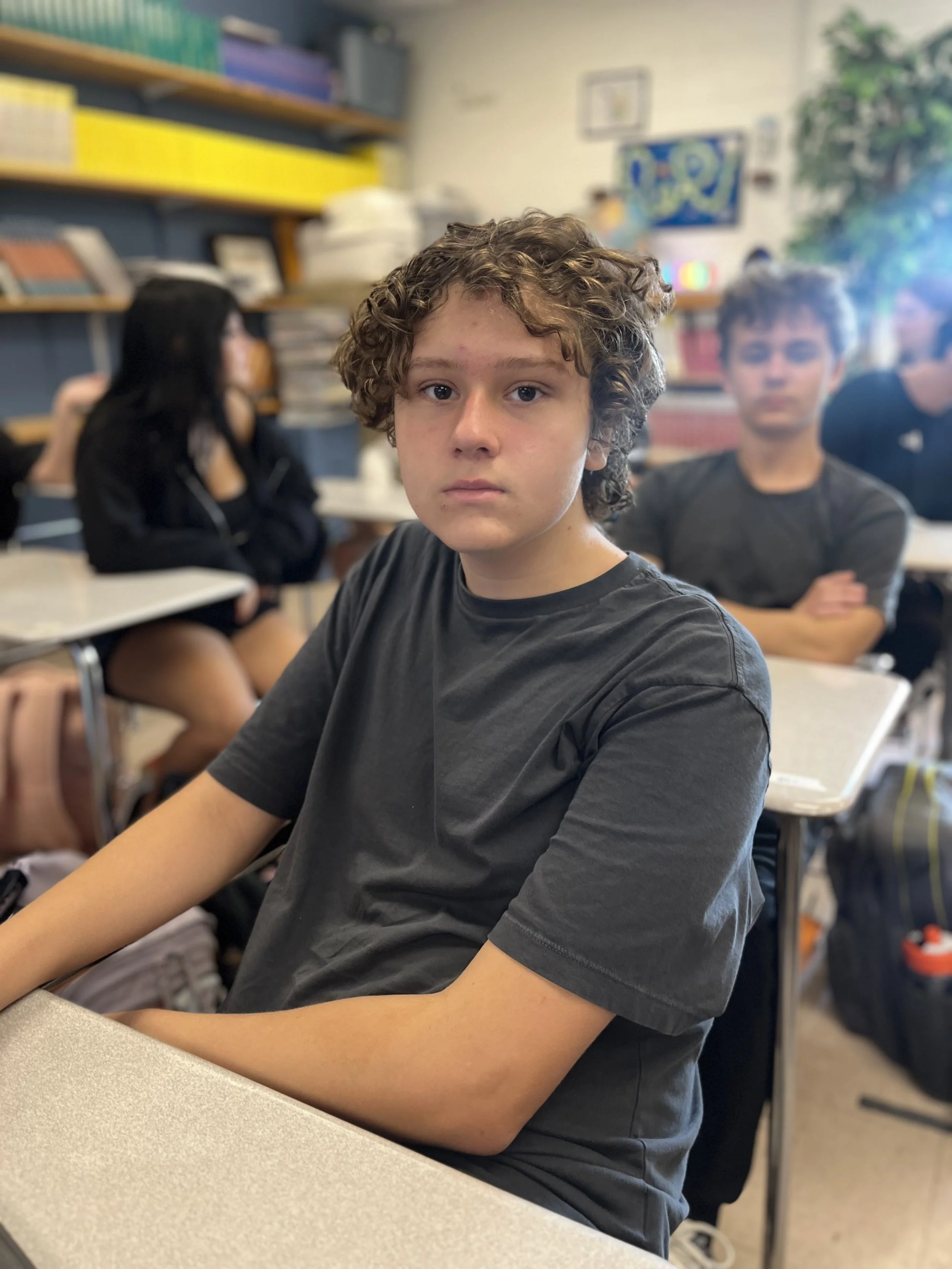A teenage girl with long brown hair, braces, and a cross necklace sitting at a desk in a classroom filled with students.