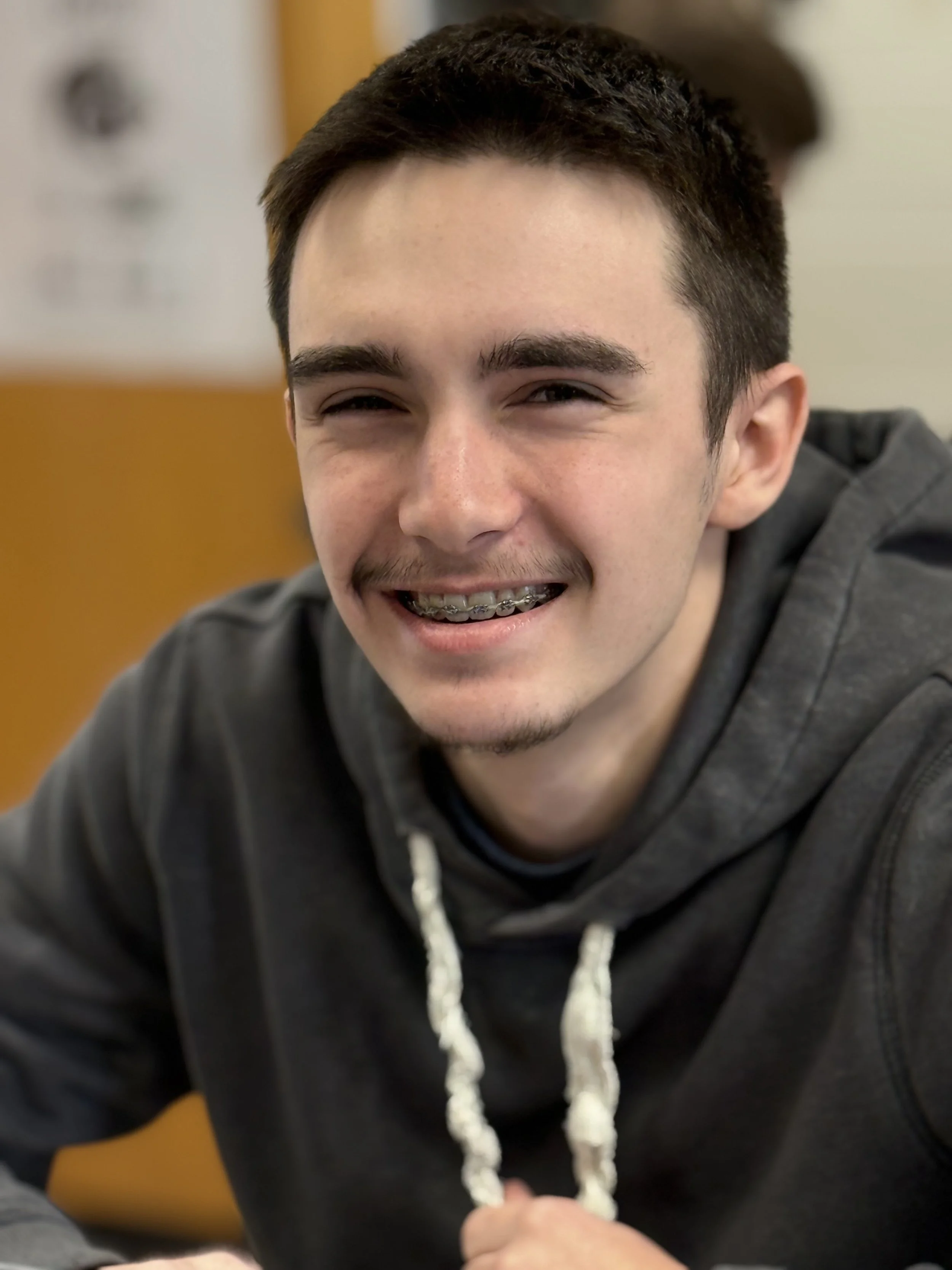 A young man smiling, wearing braces and a gray hoodie, sitting indoors with a blurred background that includes a yellow wall and some posters.