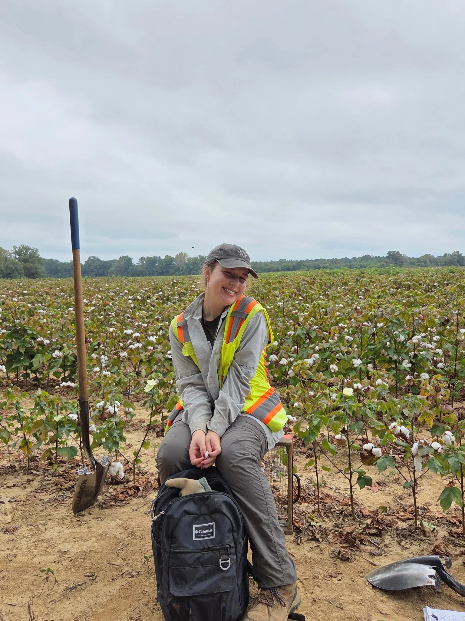 Smiling woman in safety vest and cap sitting on a stool in a cotton field, with a backpack and gardening tools, overcast sky.