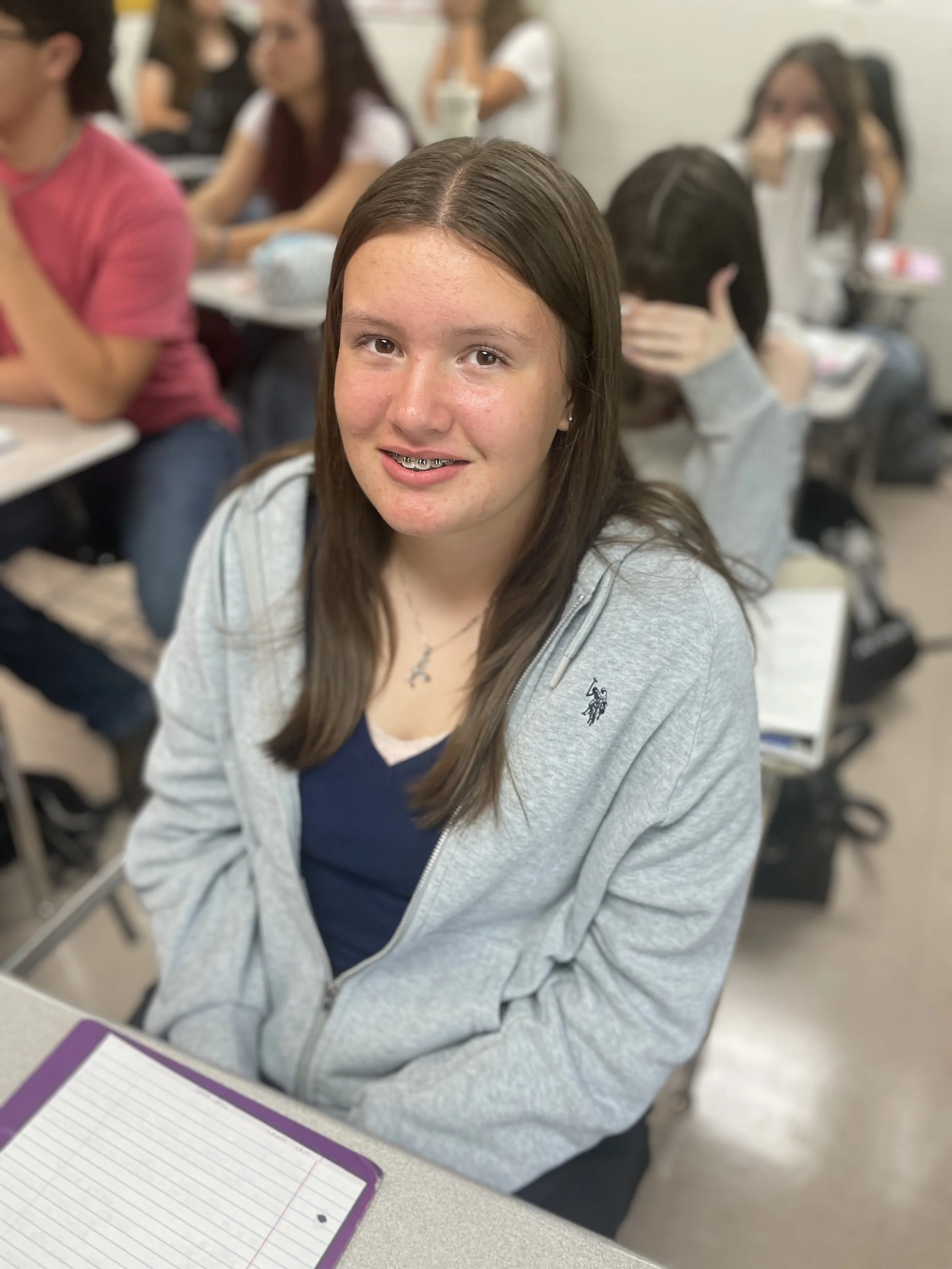 A teenage girl with long brown hair, braces, and a cross necklace sitting at a desk in a classroom filled with students.