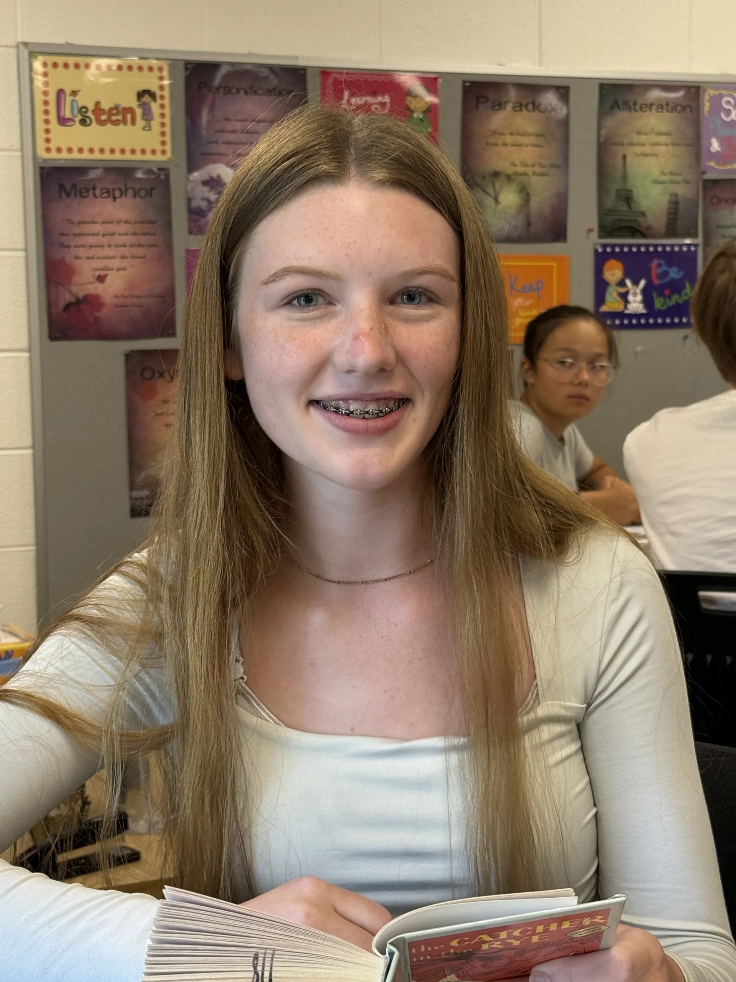 A young woman with long red hair, glasses, and braces smiling while sitting at a table in front of a red banner with a beige and gold emblem. There is a light green water bottle on the table.