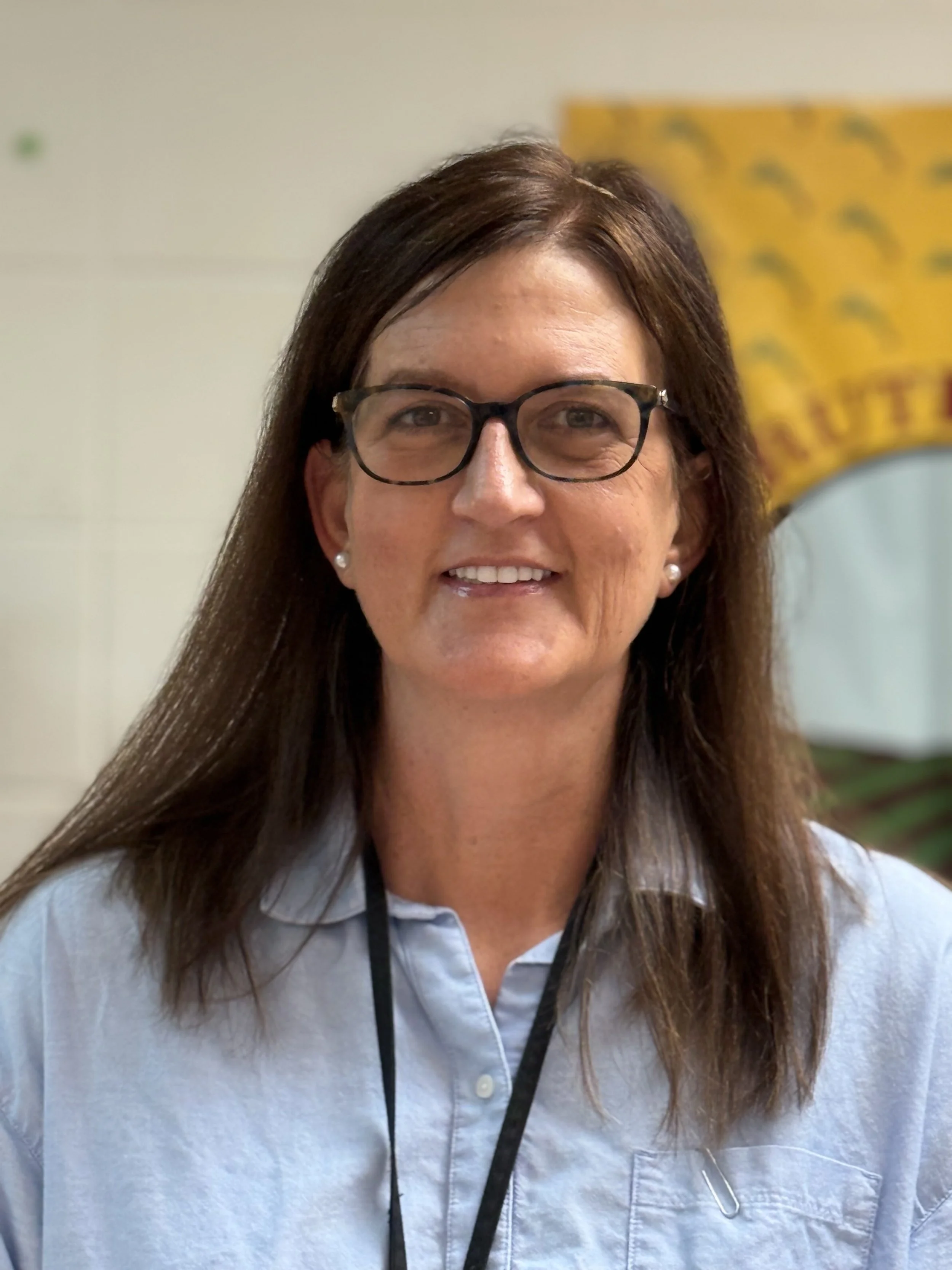 Portrait of a woman with long brown hair, wearing glasses, pearl earrings, and a light blue collared shirt, smiling.