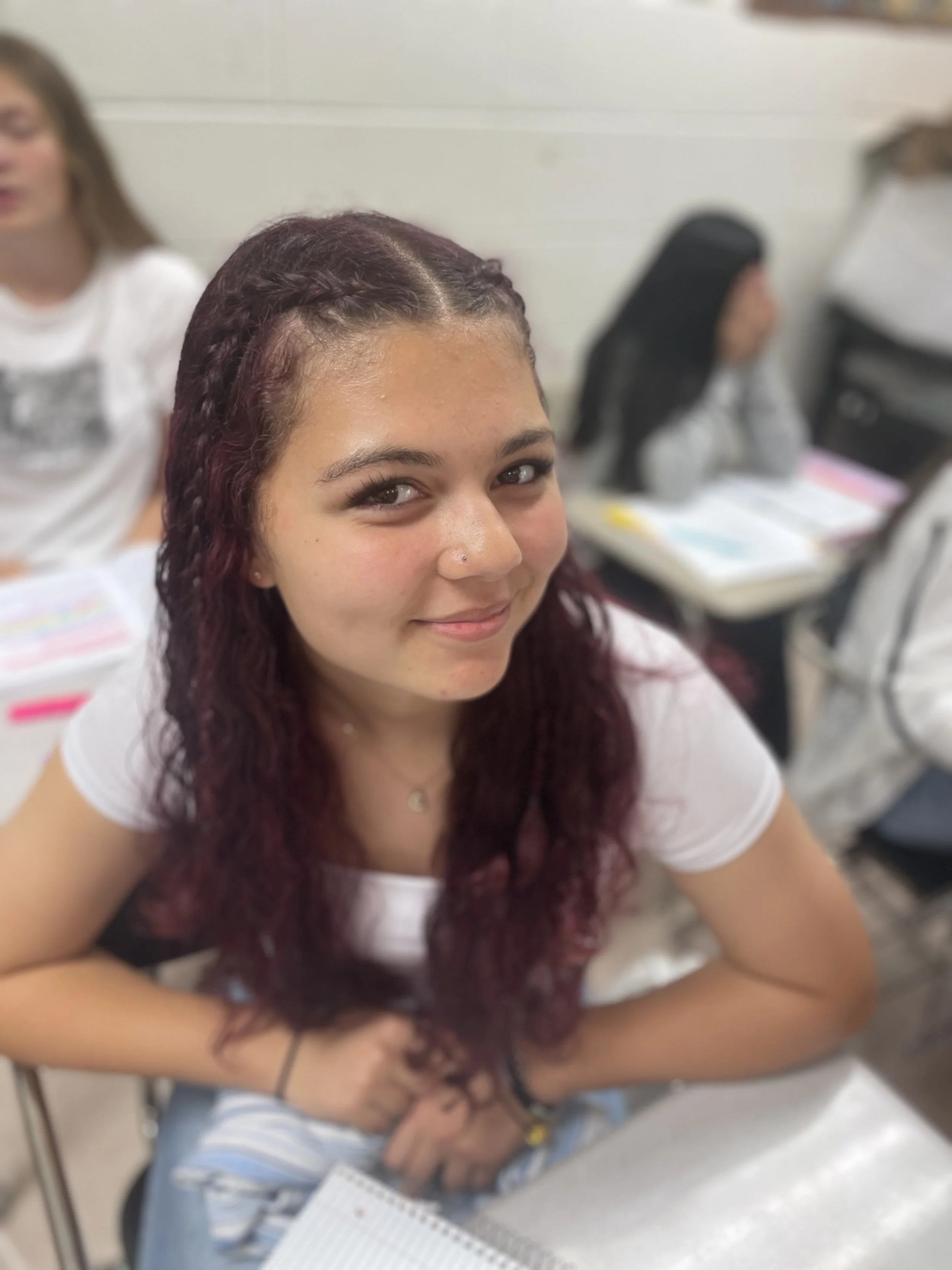 A teenage girl with long, curly, dark red hair, smiling at the camera, sitting at a desk in a classroom with other students in the background.