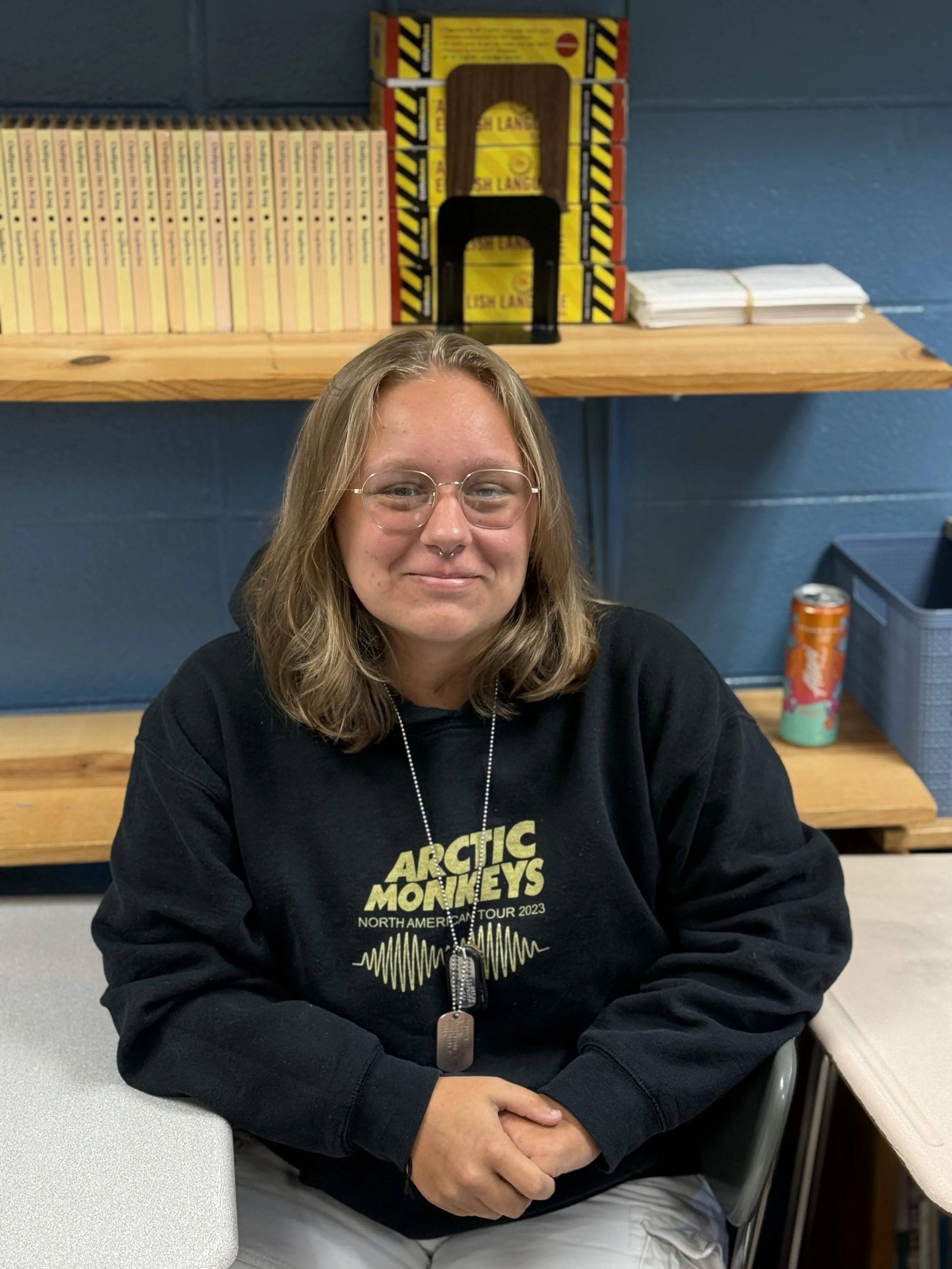 A woman with glasses and shoulder-length hair sitting at a table, wearing a black Arctic Monkeys tour sweatshirt, in a room with a wooden shelf, blue wall, notebooks, and a soda can in the background.