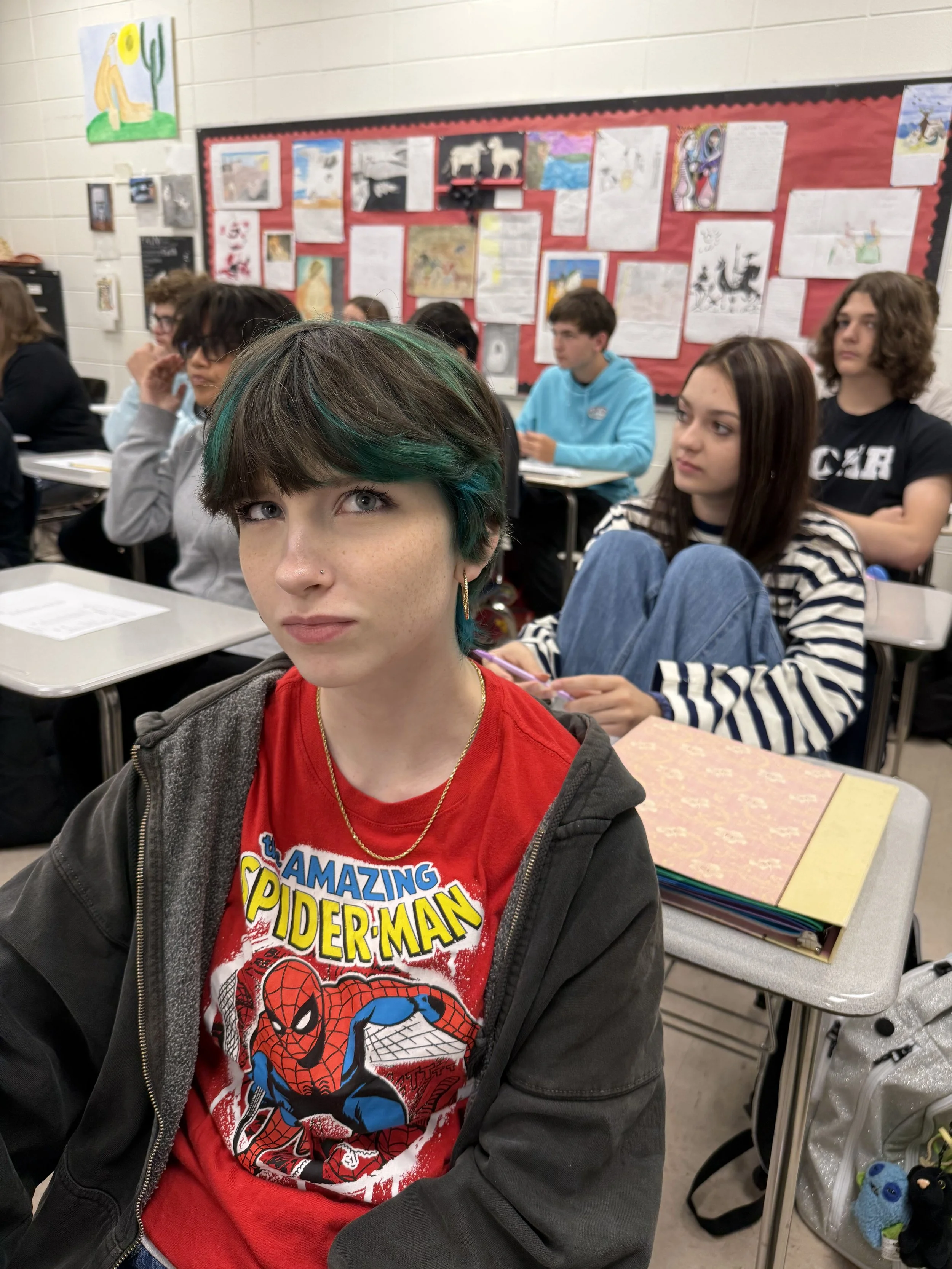A teenage girl with shoulder-length brown hair and blue eyes sitting at a school desk in a classroom, looking at the camera.