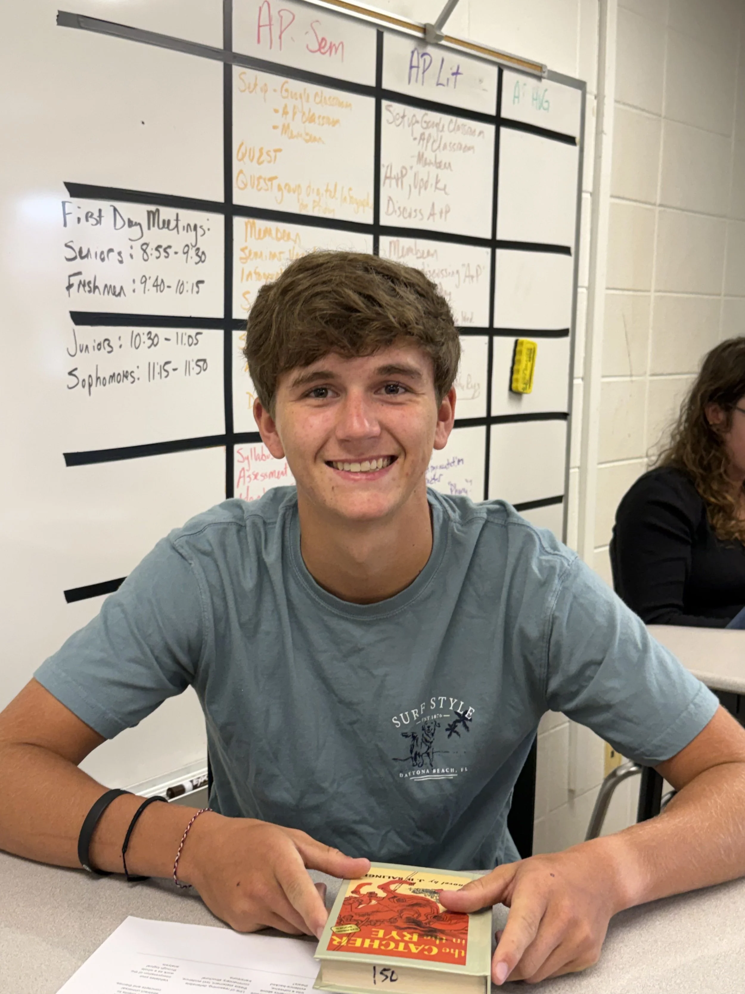 A young man sitting at a classroom desk, holding a yellow pencil, with papers in front of him, wearing a white t-shirt that says "Ridge Land Honors Academy" and a smartwatch on his left wrist.