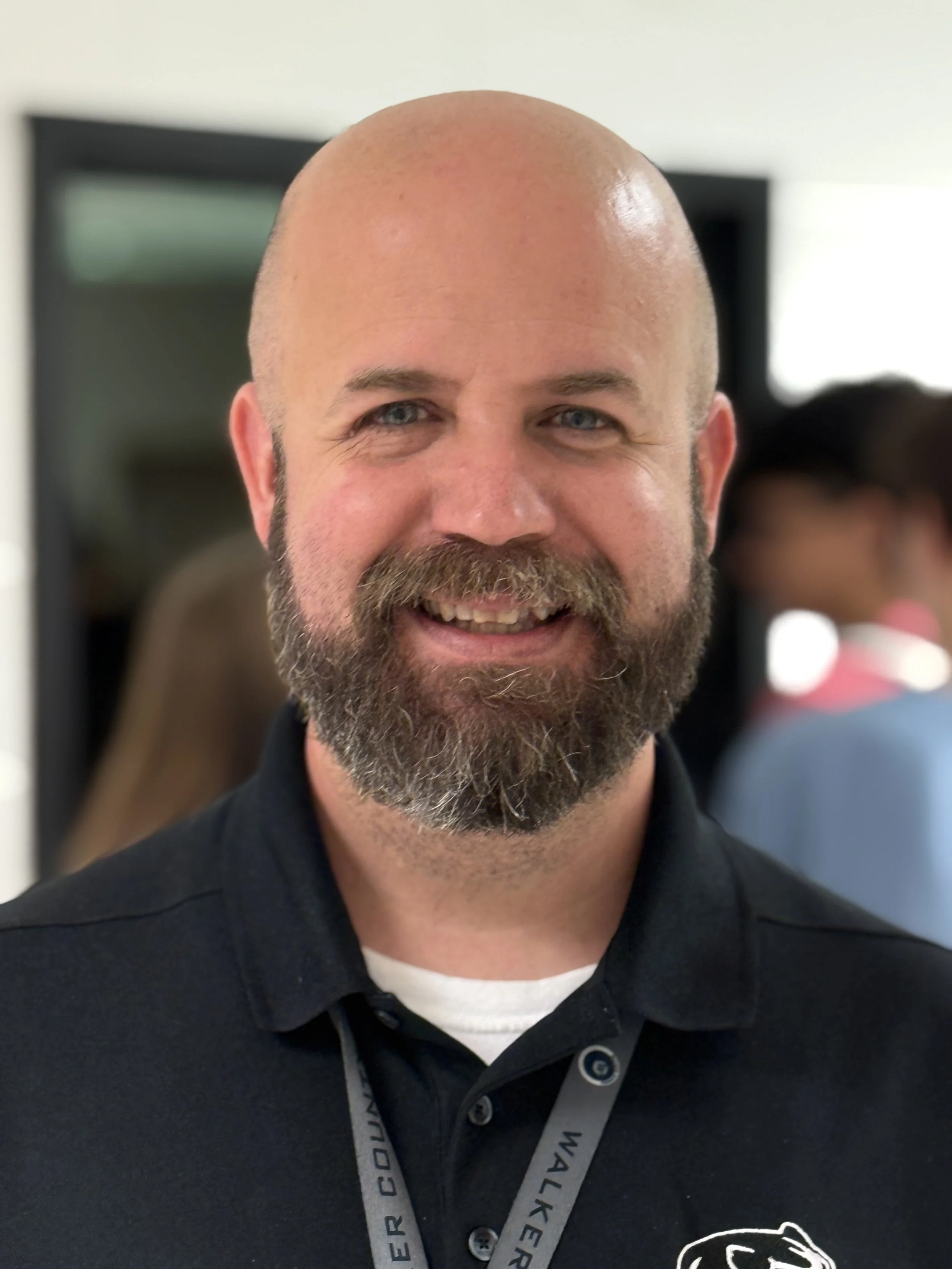 A smiling man with a beard and shaved head, wearing a black polo shirt with a lanyard around his neck, standing indoors with a blurred background of people and a mirror.