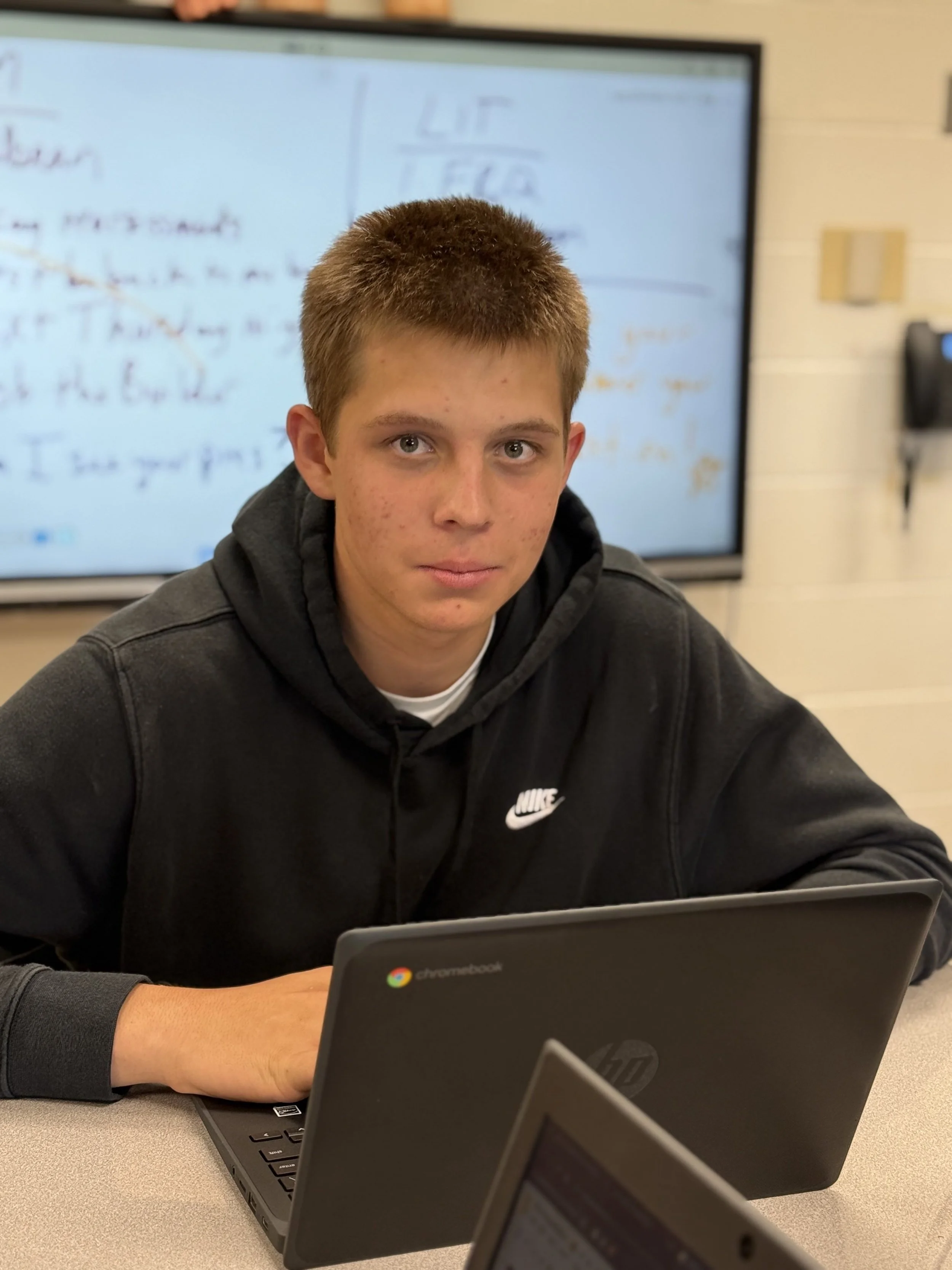 A teenage boy with short brown hair and light skin, wearing a black Nike hoodie, sitting at a desk with a Chromebook in front of him, in a classroom setting with a whiteboard in the background.