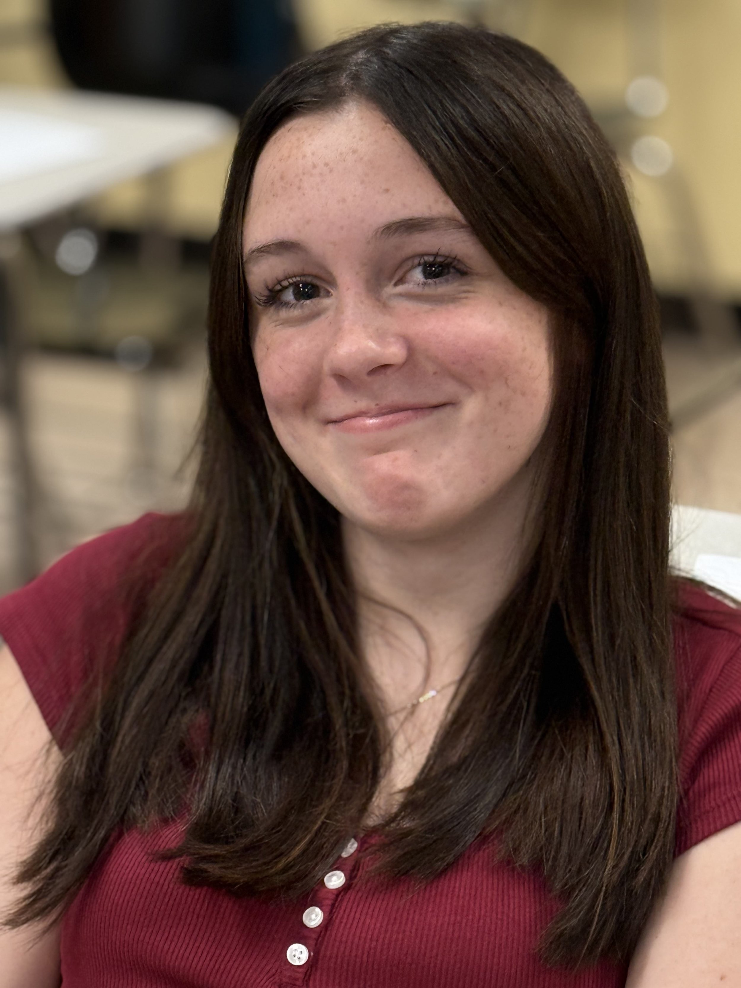 A young woman with long dark brown hair, fair skin, and freckles, smiling and looking at the camera.
