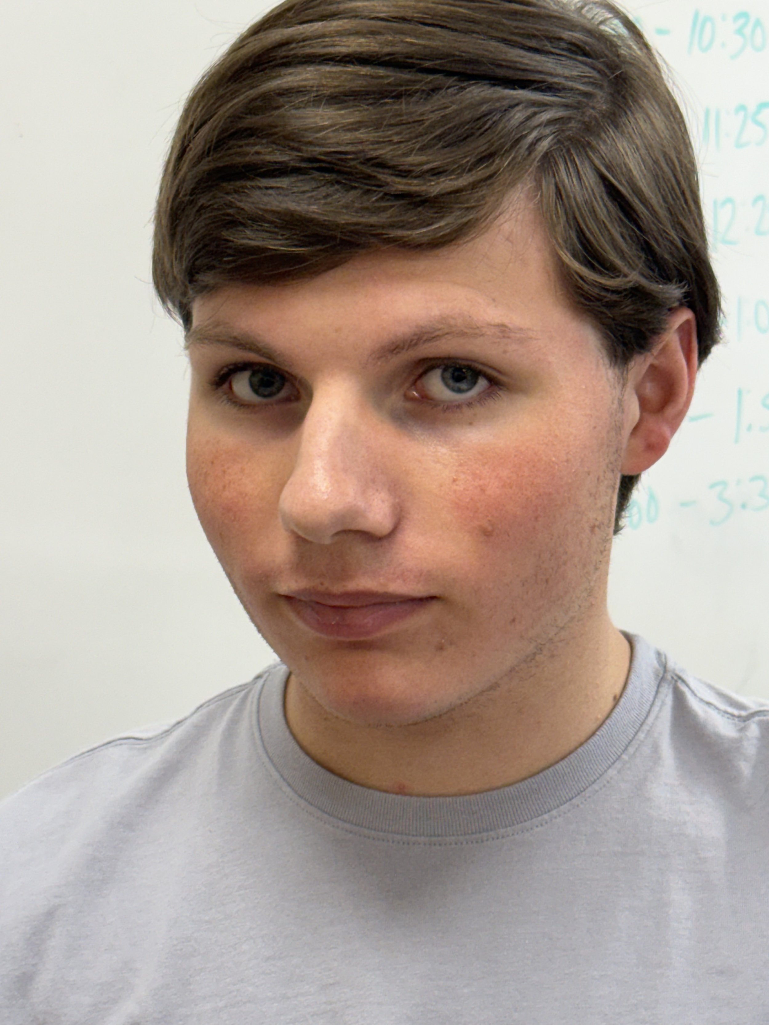 A teenage boy with brown hair and blue eyes is sitting at a desk in a classroom, holding a yellow pencil, with a notebook open in front of him. There is a blurred classroom presentation in the background.