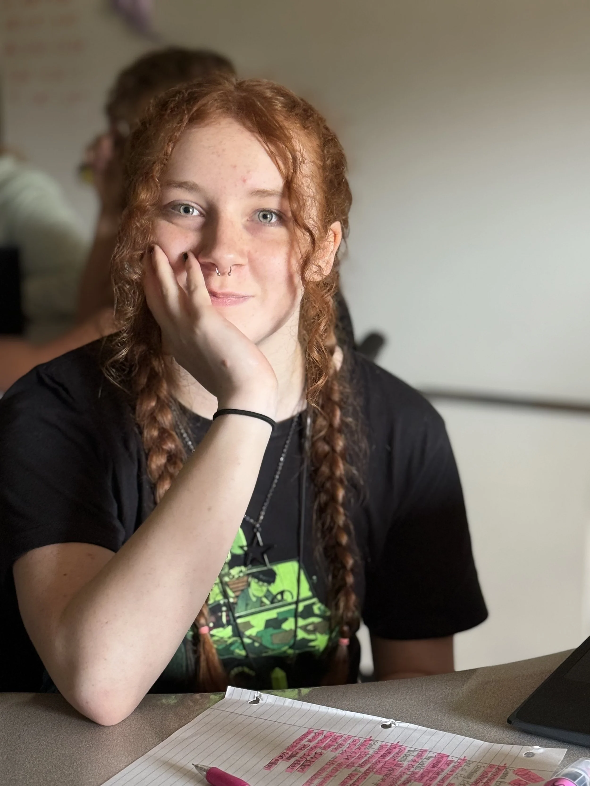 A young woman with red hair in braids, light skin, and a septum piercing, sitting at a desk with lined paper and pink highlighter, resting her chin on her hand.