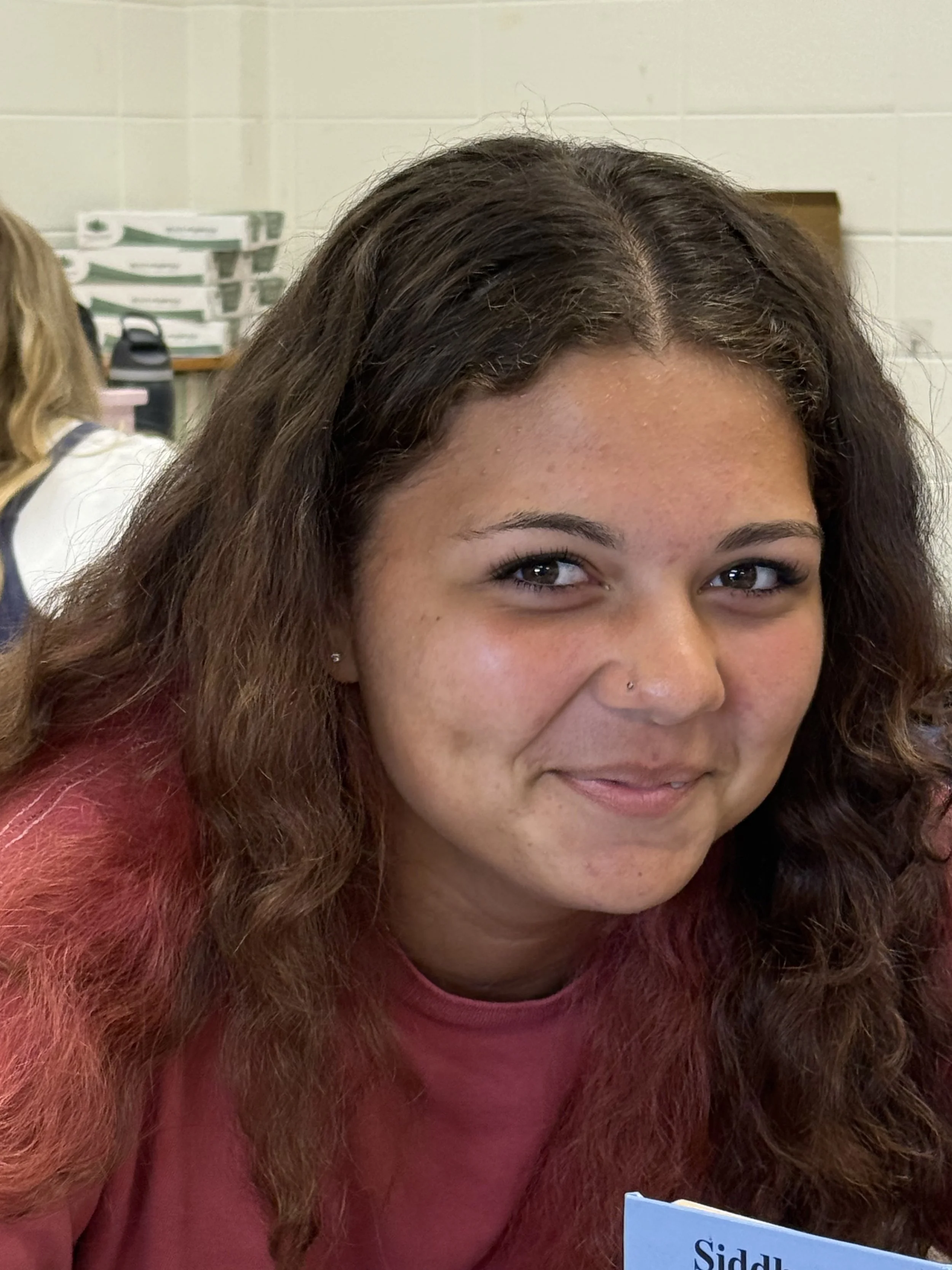 A teenage girl with long, curly, dark red hair, smiling at the camera, sitting at a desk in a classroom with other students in the background.