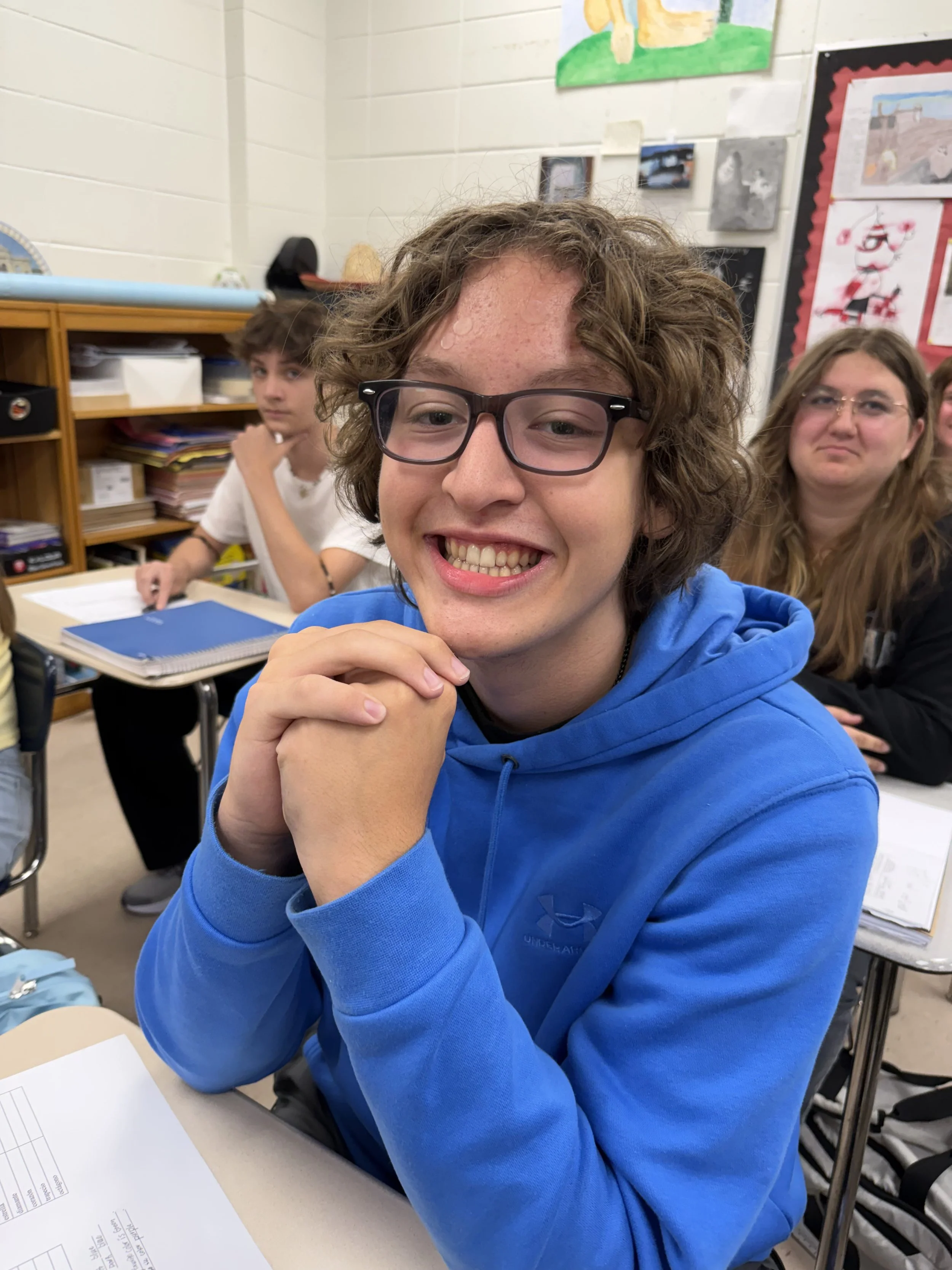 Young man with curly hair and glasses sitting in a classroom, holding a green striped straw, with other students and a computer in the background.