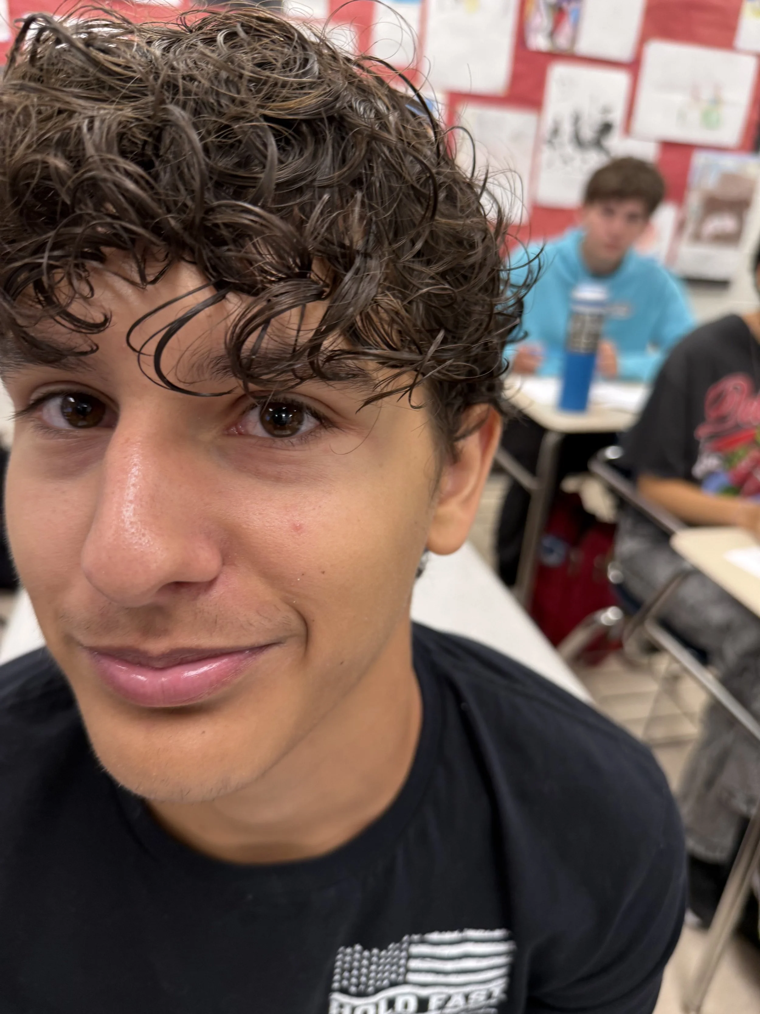 A boy with curly hair and a beige hoodie sitting at a desk in a classroom, with other students in the background.