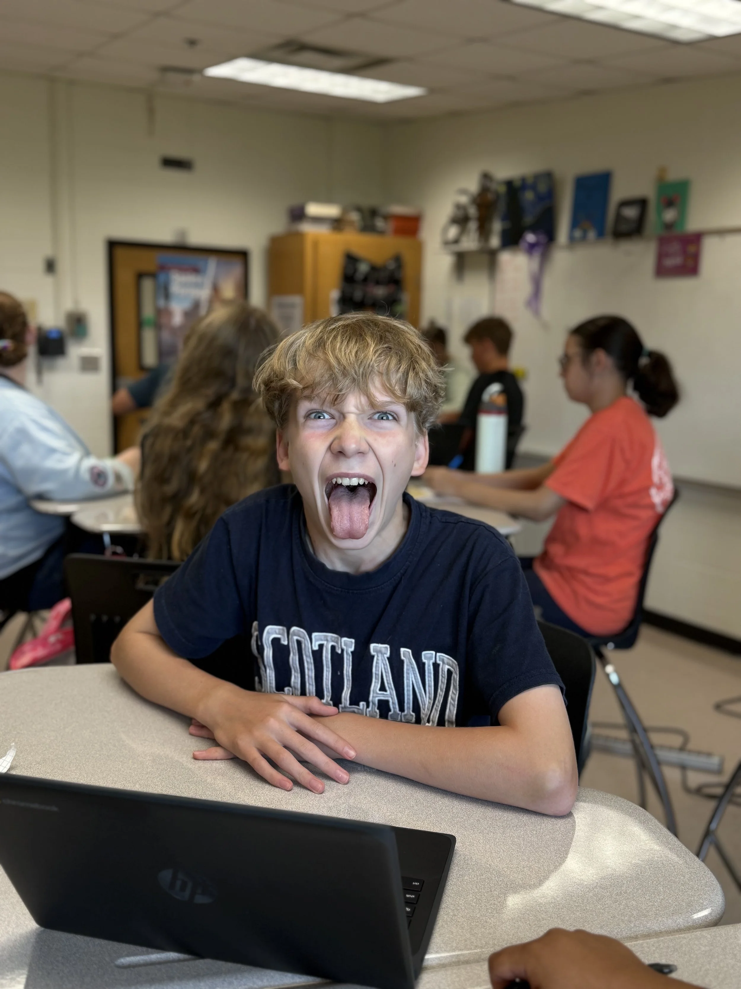 A young boy with messy blond hair making a silly face, sticking his tongue out and opening his mouth wide, sitting at a desk in a classroom with other students in the background.