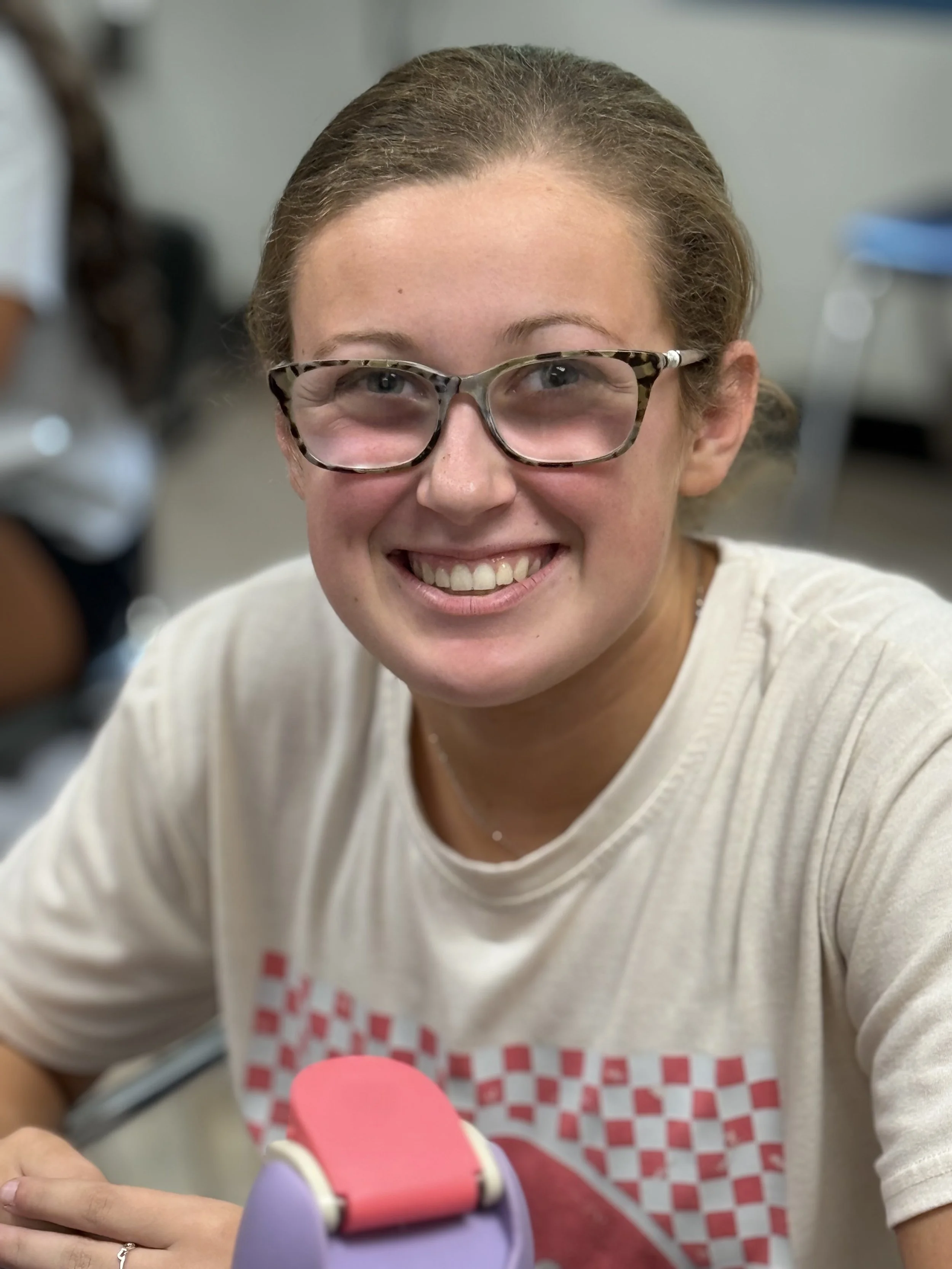A young woman with glasses smiling at the camera, sitting at a table in a classroom or office setting.