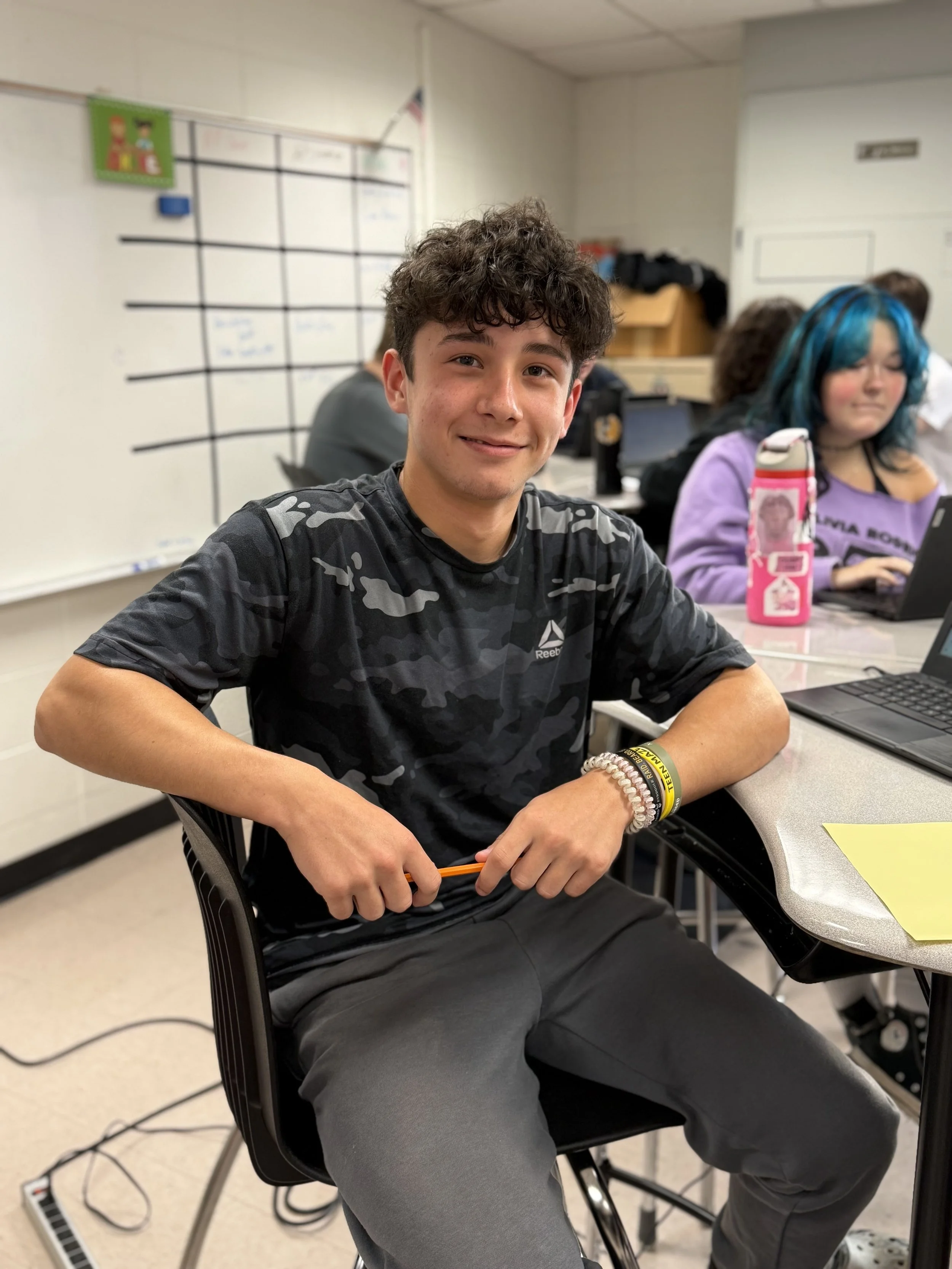 A young man with curly brown hair sitting at a desk in a classroom, smiling at the camera, wearing a black camouflage patterned Reebok t-shirt, watching a girl with blue hair working on her laptop in the background.