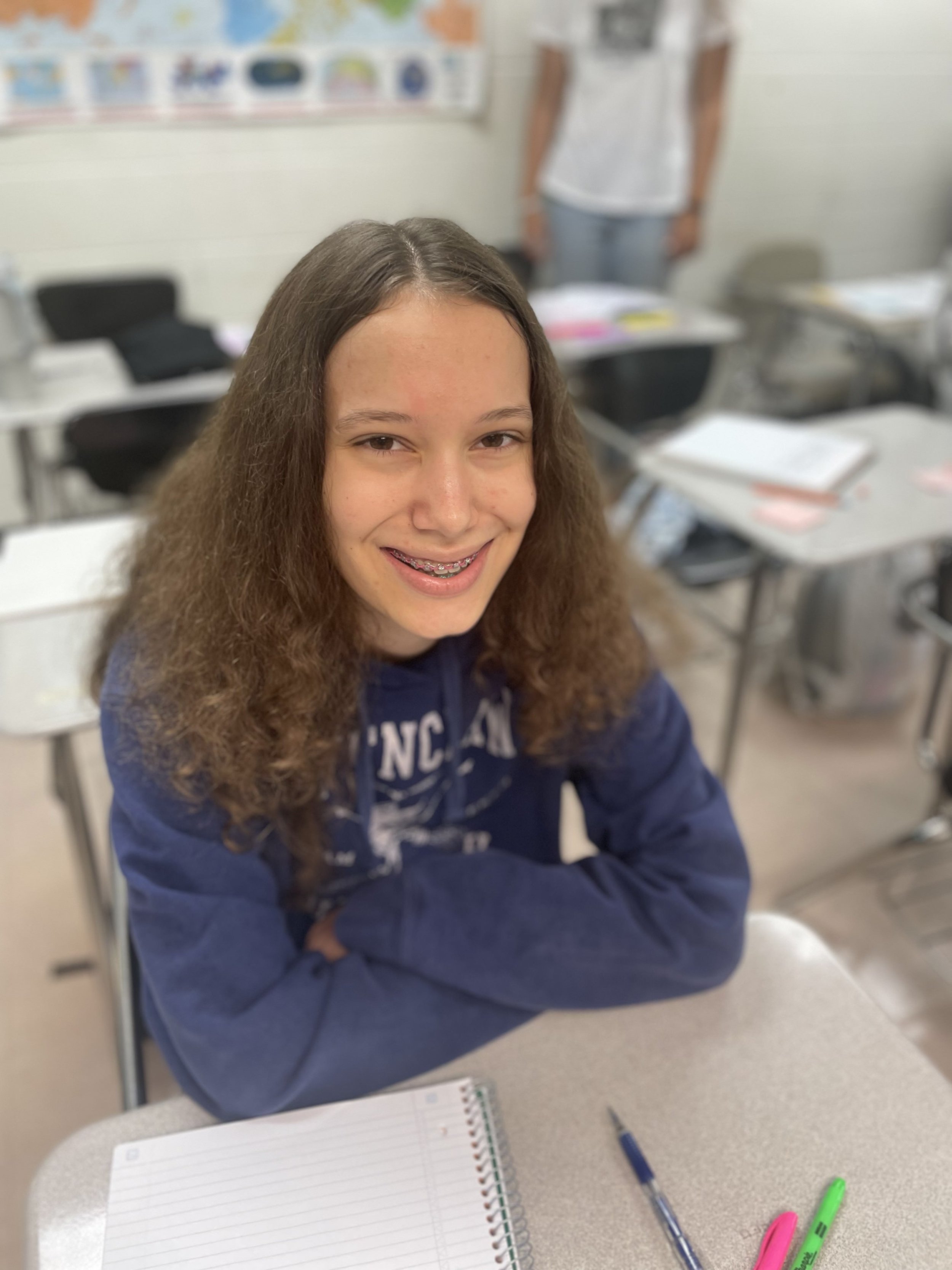 A smiling girl with braces sitting at a desk in a classroom, wearing a blue hoodie with her arms crossed on the table. There are notebooks and pens on the desk.