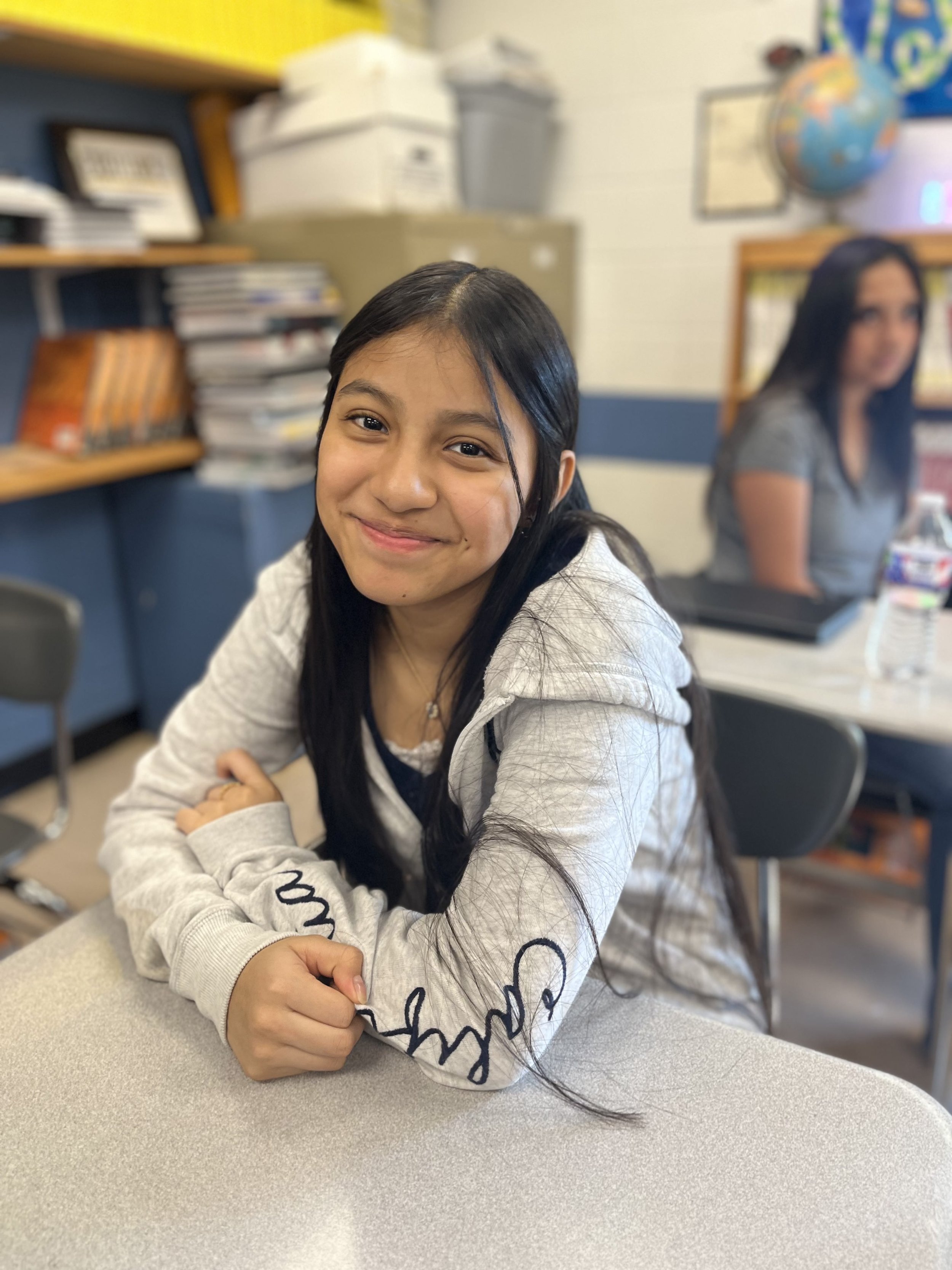 Young woman with glasses and brown hair making a peace sign near her face, sitting at a desk with water bottles in front, against a whiteboard background.