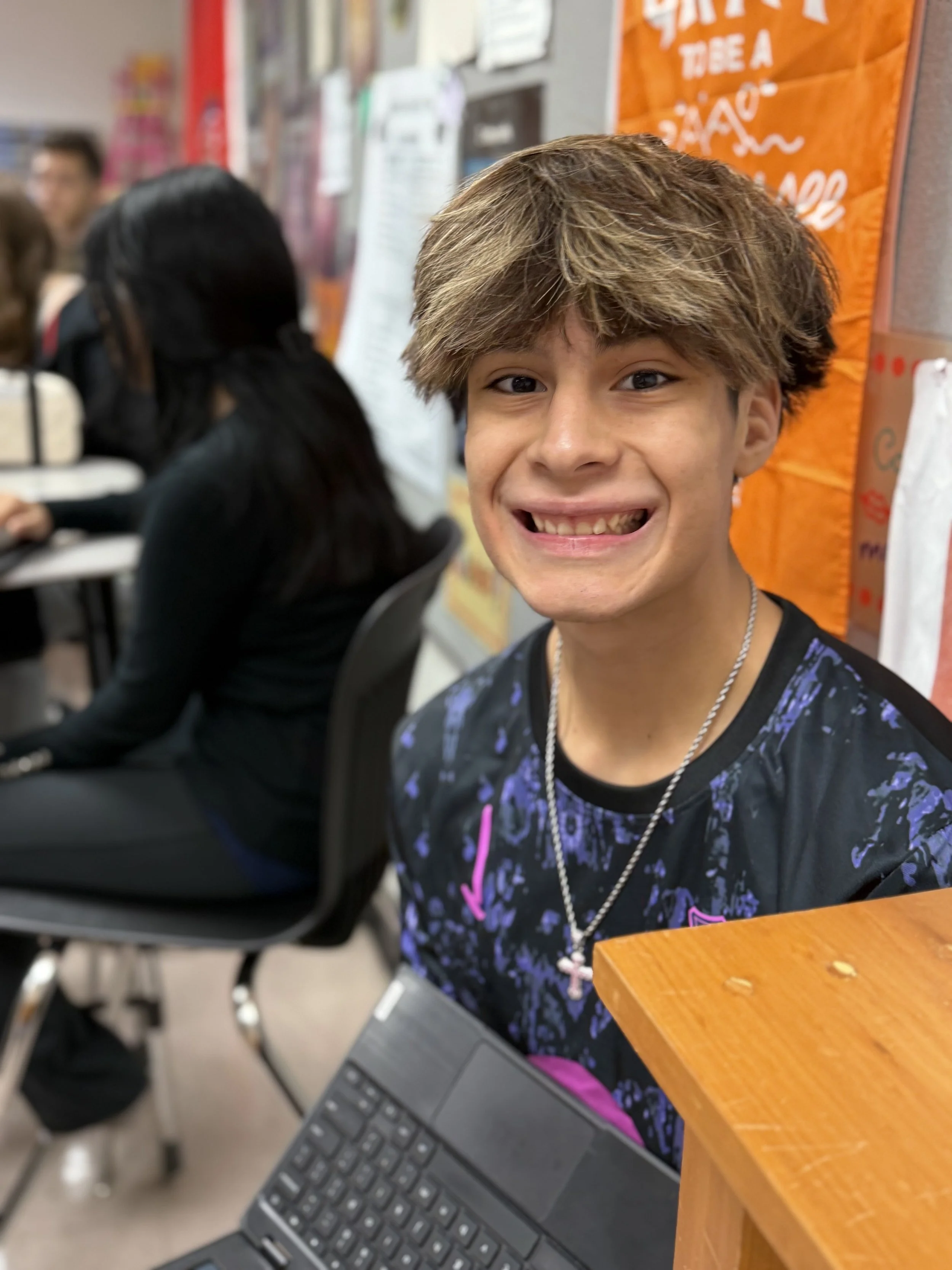 A smiling teenage boy with light brown, tousled hair and a cross necklace, sitting in a classroom with others in the background, at a desk with a laptop.