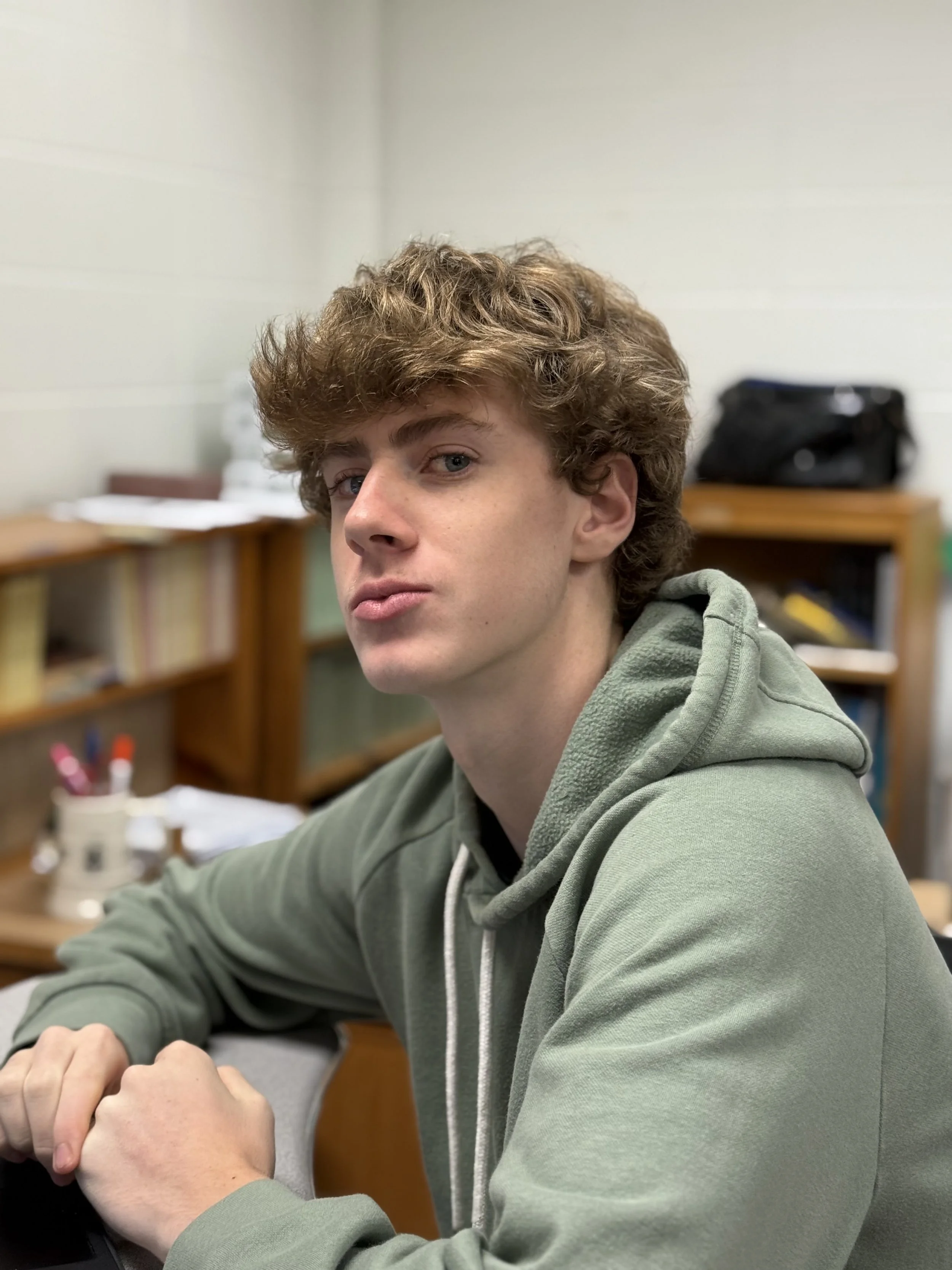 A young man with curly, light brown hair and blue eyes sitting at a desk with a serious expression, wearing a green hoodie, in a room with bookshelves and office supplies.
