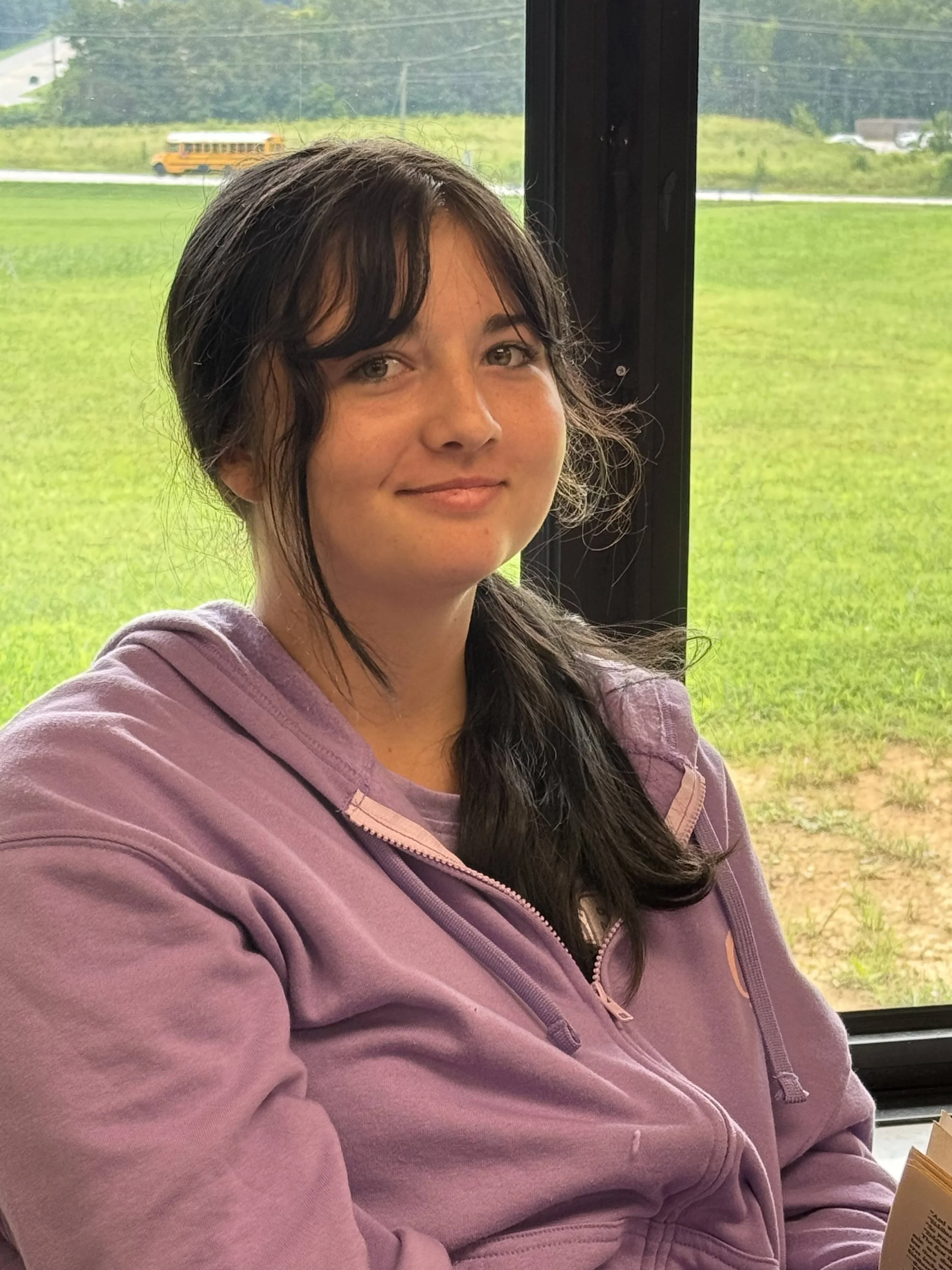 A teenage girl with long brown hair and blue eyes smiling softly, with a blurred person in the background.