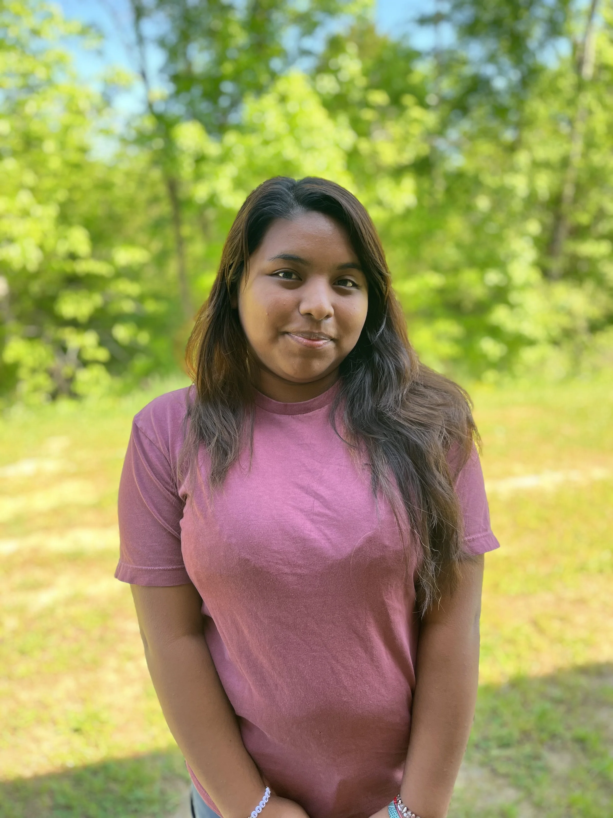 A woman with long black hair smiling and wearing a green shirt, sitting indoors with a colorful blurred background.