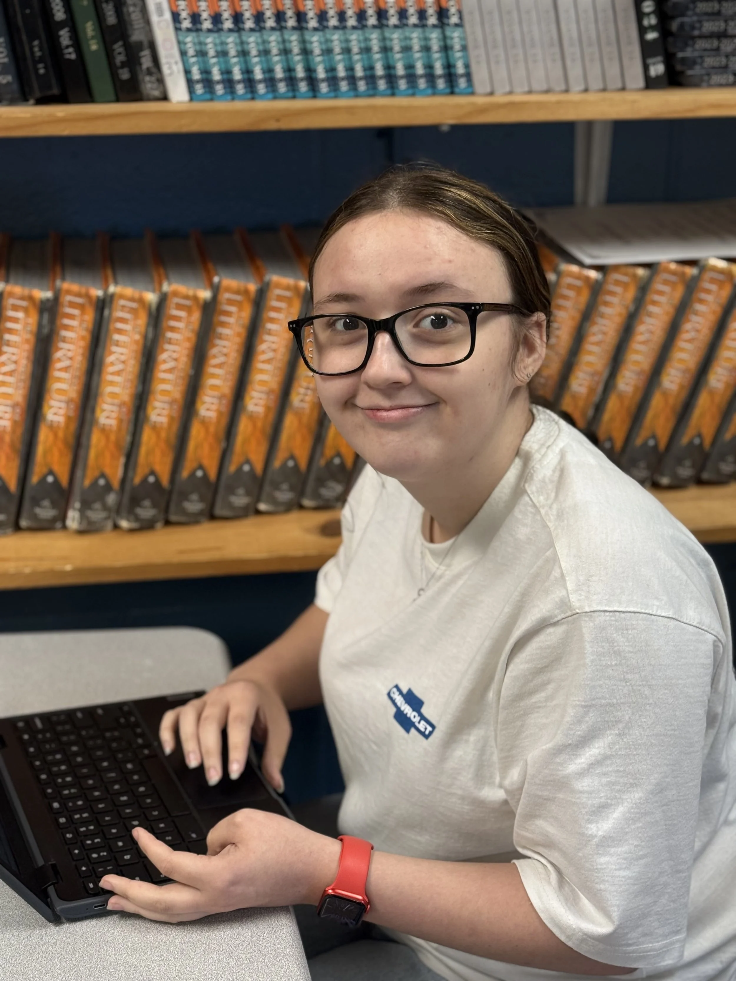 A young woman with glasses and a white shirt sitting at a desk with a laptop, in a library or bookstore, with shelves of books behind her.
