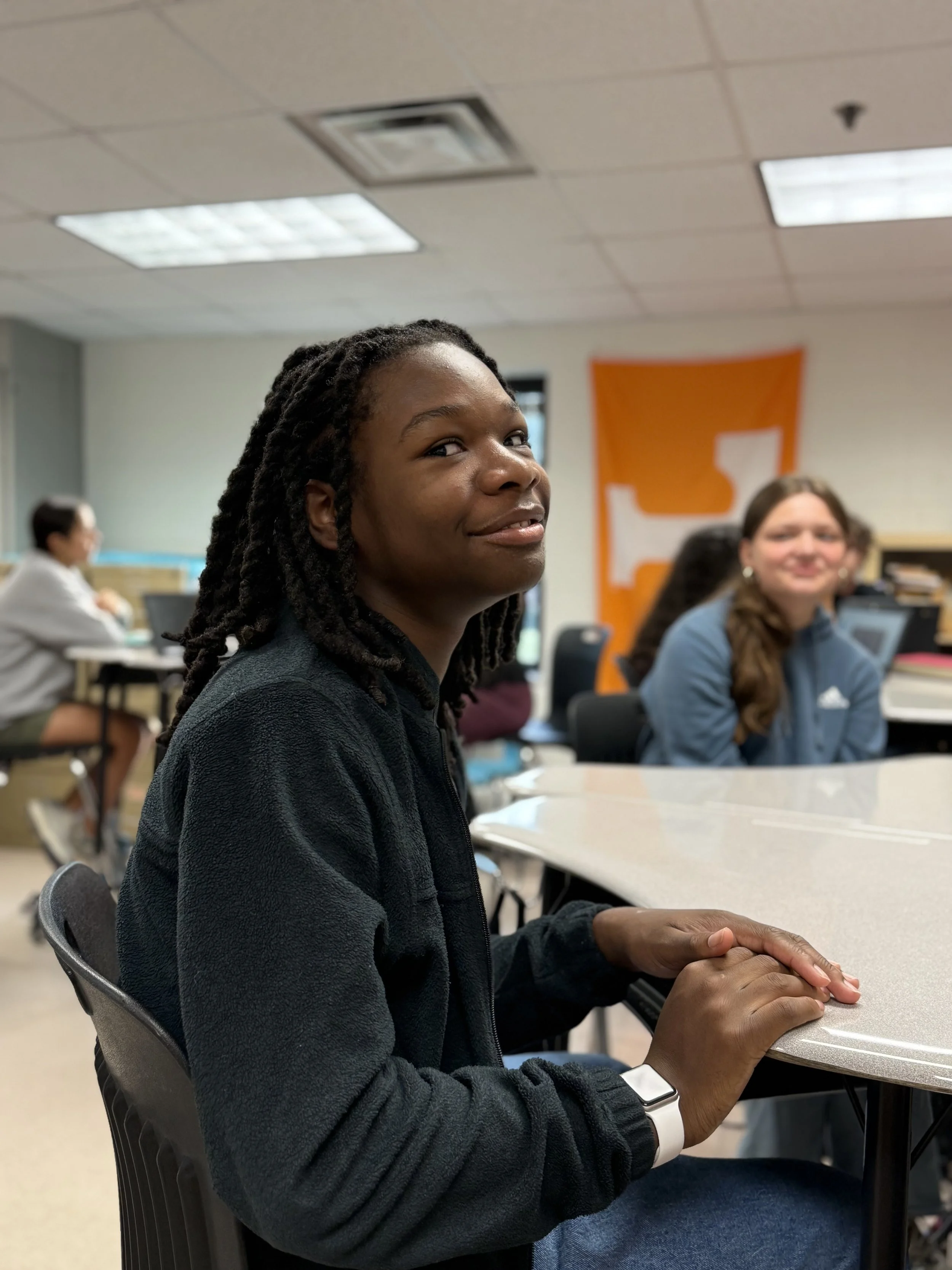 A young woman with braided hair sitting at a table in a classroom, looking at the camera with a slight smile. There are other students and an orange and white banner with a large 'T' in the background.