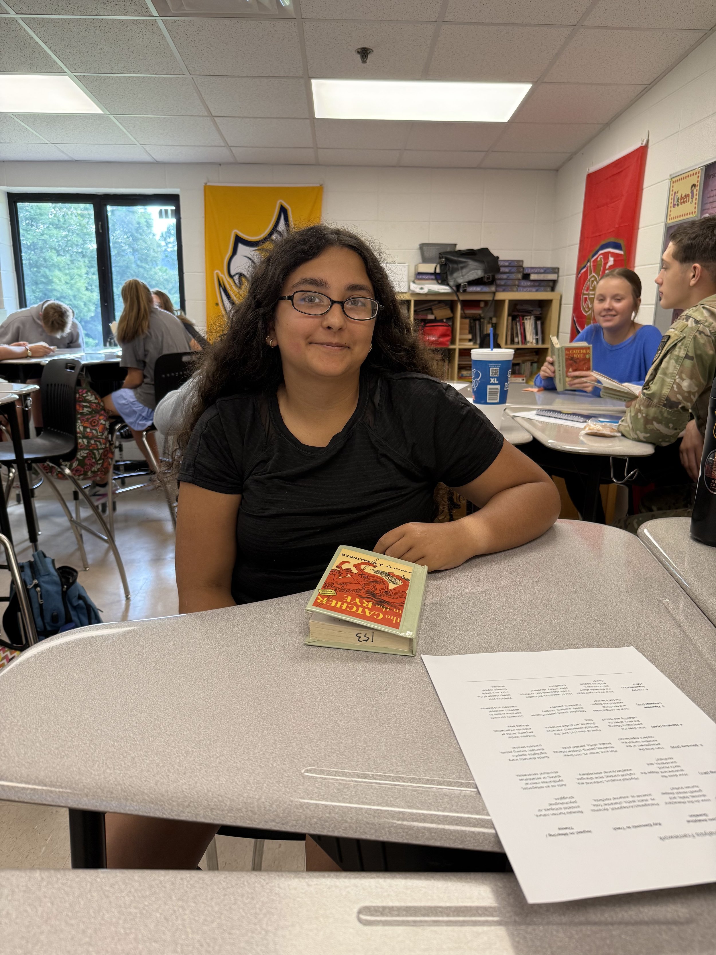 A young woman with glasses and curly hair, smiling at the camera, sitting at a desk in a classroom with other students and large windows in the background.