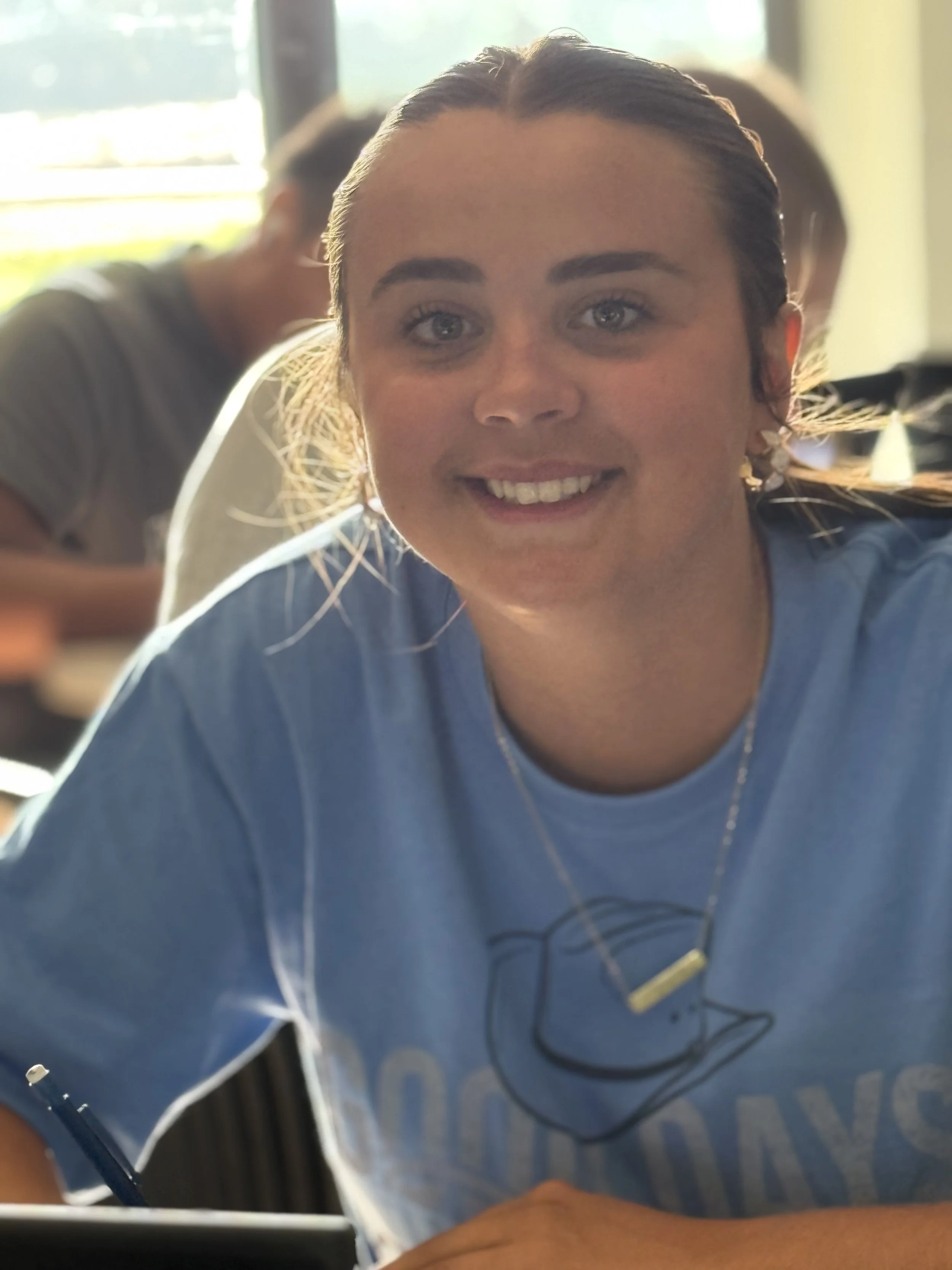 A smiling young woman with braided hair, wearing a blue t-shirt with a graphic and a necklace, sitting at a table in a well-lit room.
