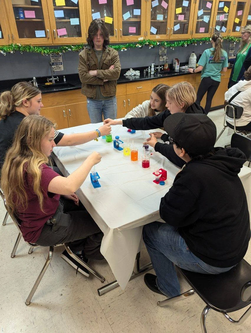 Group of students doing a science experiment in a classroom. Five students sit around a table with colorful liquids and a few science tools. Two students stand in the background.