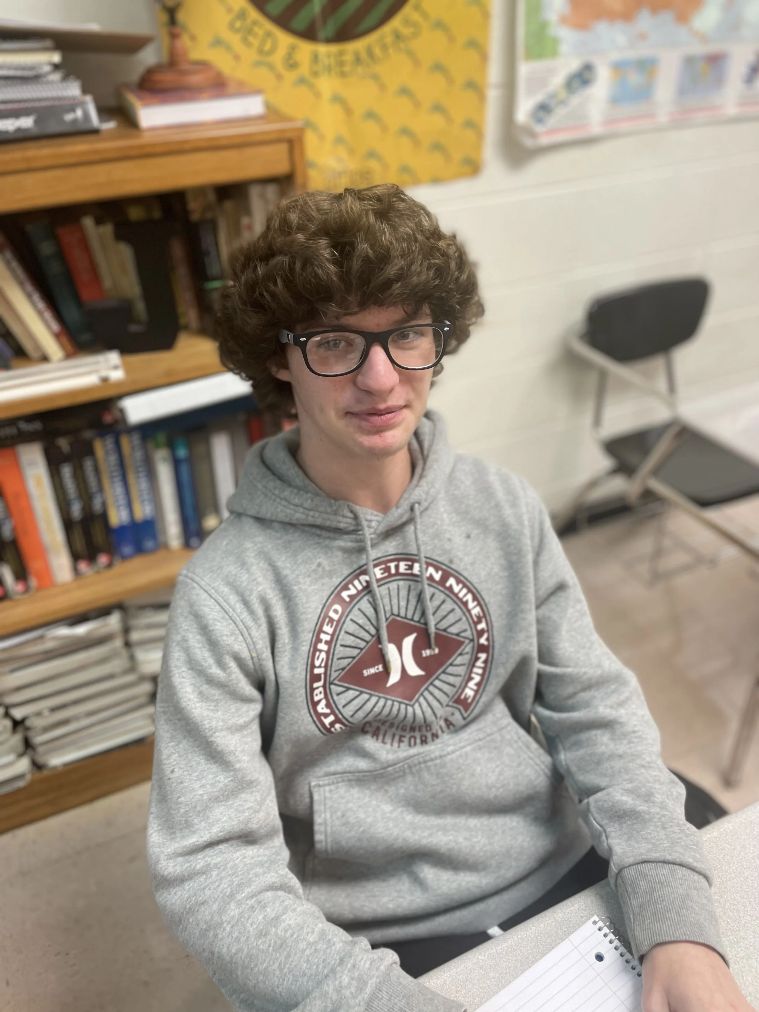 A young man with curly brown hair and glasses sitting in a classroom, wearing a gray hoodie with a circular logo on it, with bookshelves and educational posters in the background.