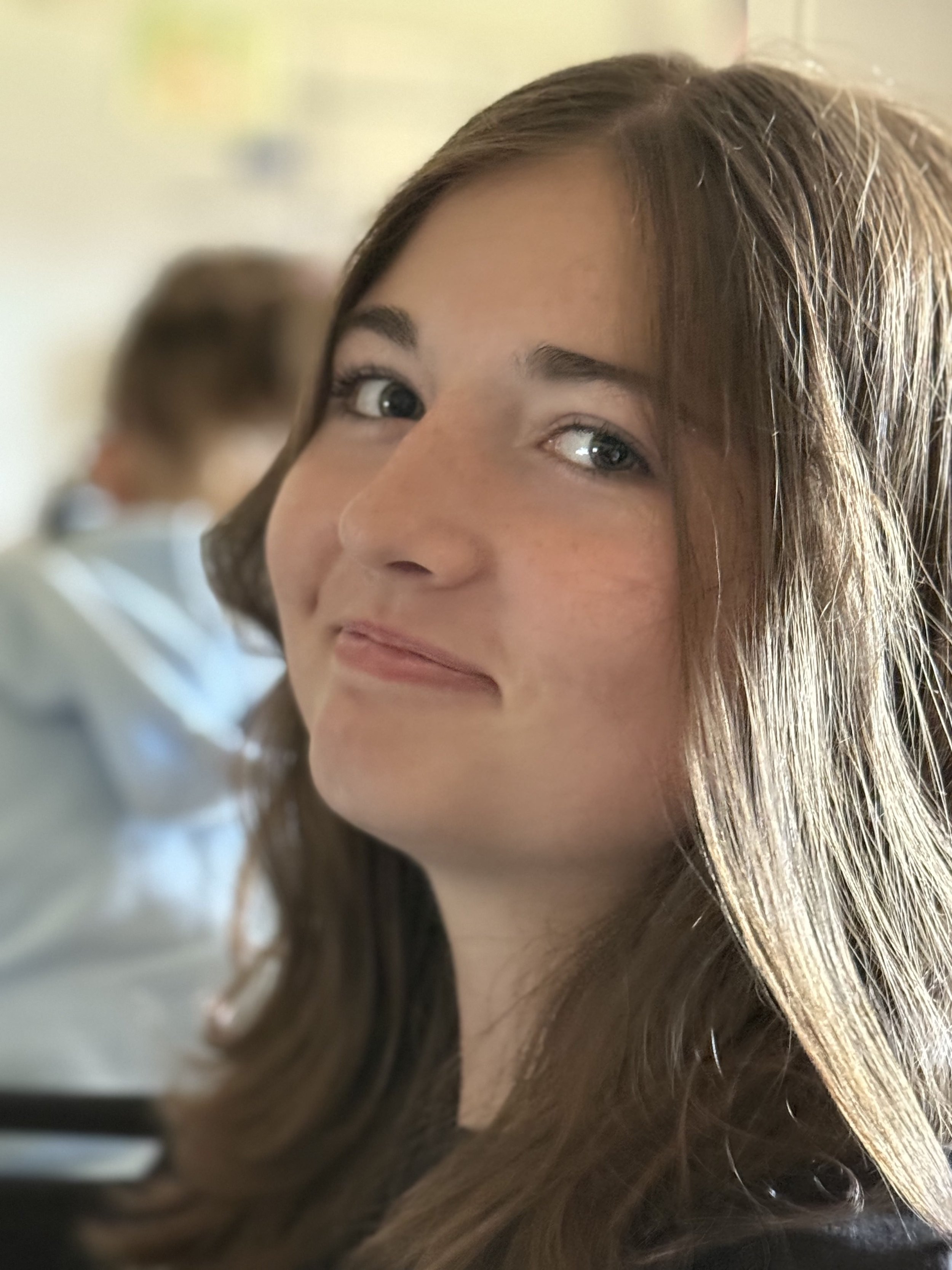 A teenage girl with long brown hair and blue eyes smiling softly, with a blurred person in the background.