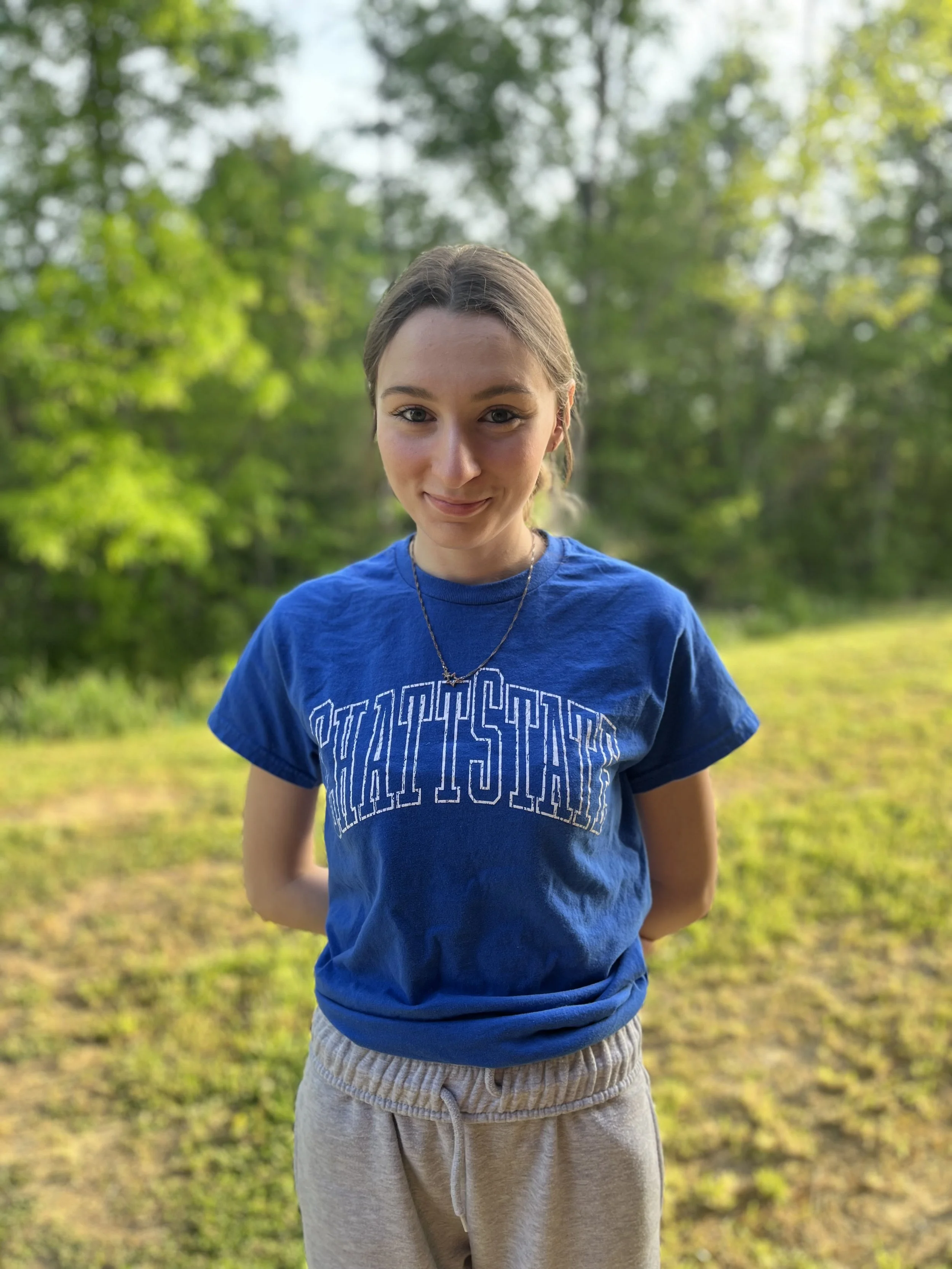 A young woman with shoulder-length light brown hair smiling, wearing a black top with lace shoulder detail, earrings, and a necklace, against a blurred colorful background.
