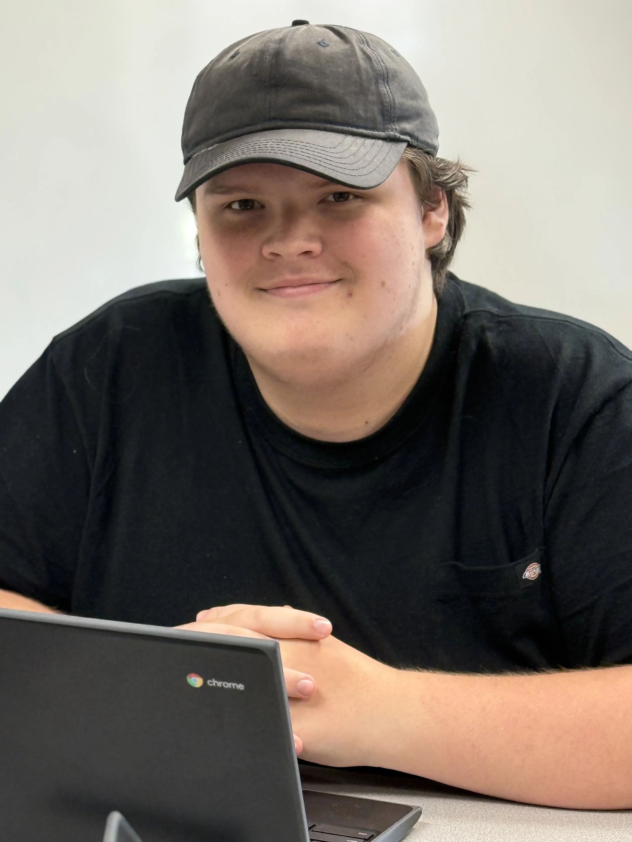 A young man wearing a gray baseball cap and black t-shirt, sitting at a table with a laptop.