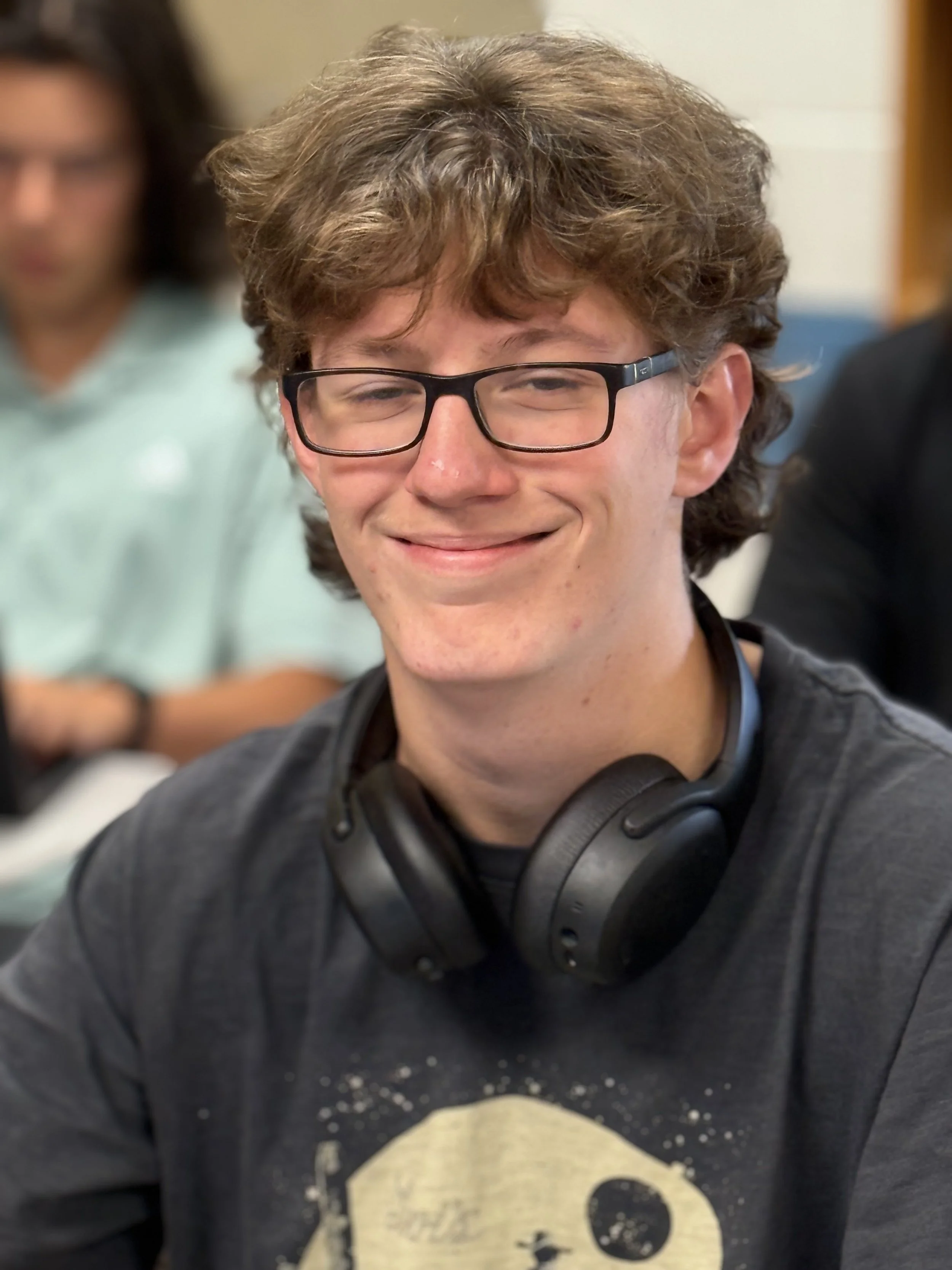 Young man with curly hair and glasses smiling, wearing a black t-shirt with a skull design, and headphones around his neck, in a classroom setting.