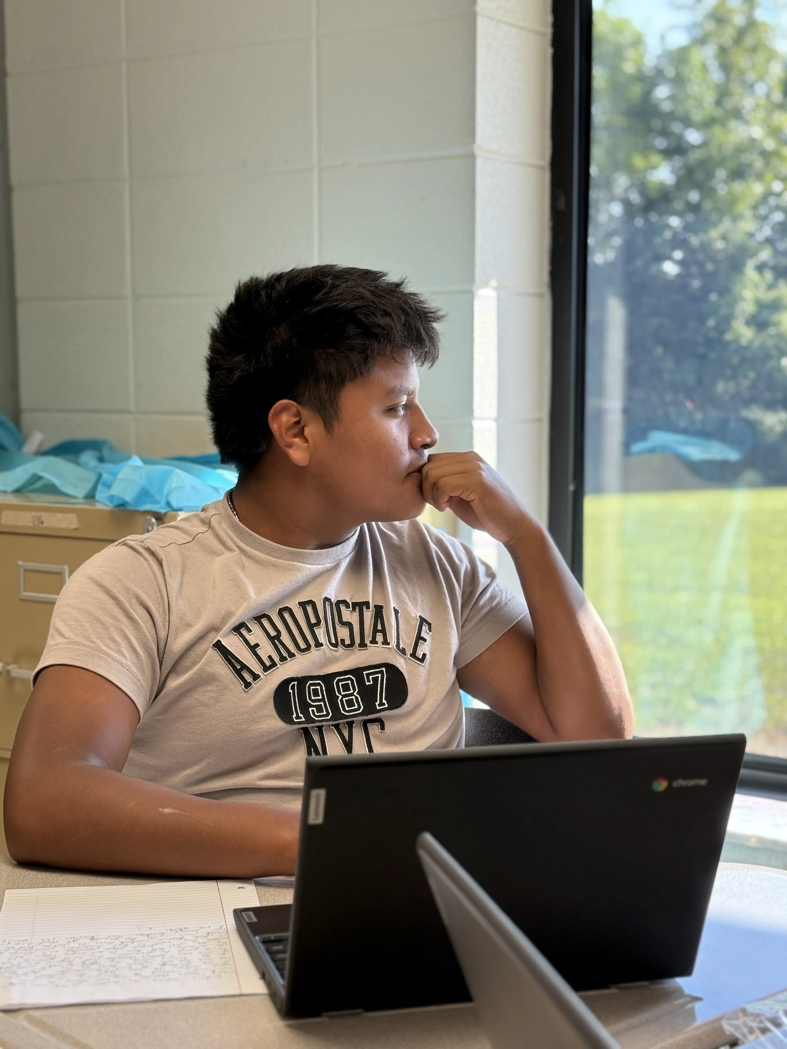A young man with short dark hair sitting at a desk, looking thoughtfully out a window. He is wearing a gray Aeropostale t-shirt and has a notebook with handwritten notes and a closed Chromebook in front of him.