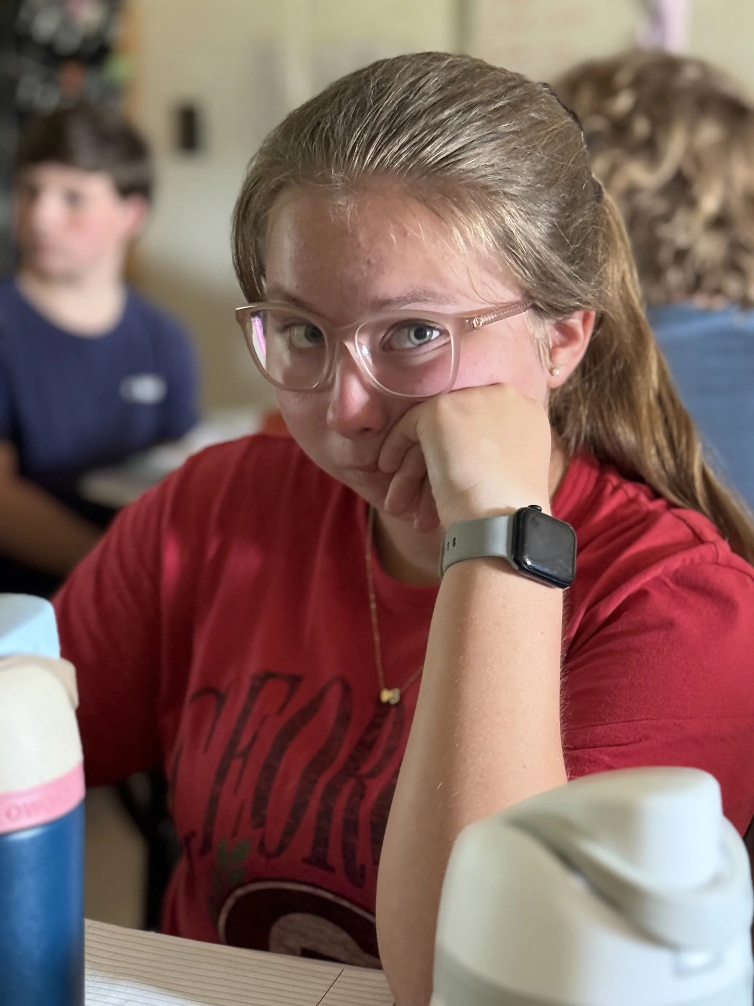 A young girl wearing glasses and a red shirt sitting at a table, resting her chin on her hand, looking directly at the camera. She has a smartwatch on her wrist and is in a room with other people in the background.